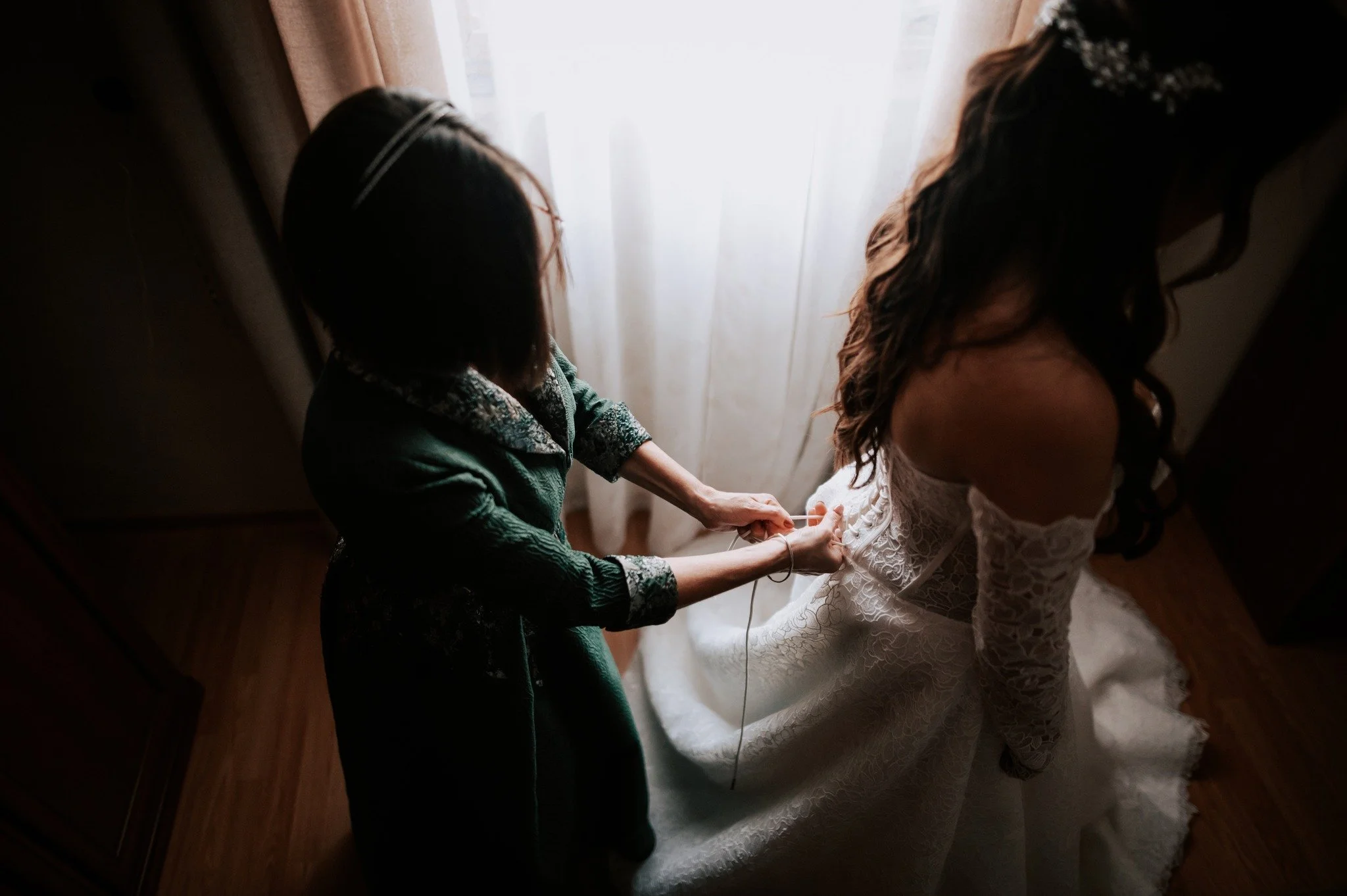 A woman in a green robe is tying the laces on the back of a bride's white lace wedding dress while standing by a window with sheer curtains, with a wooden floor beneath them.