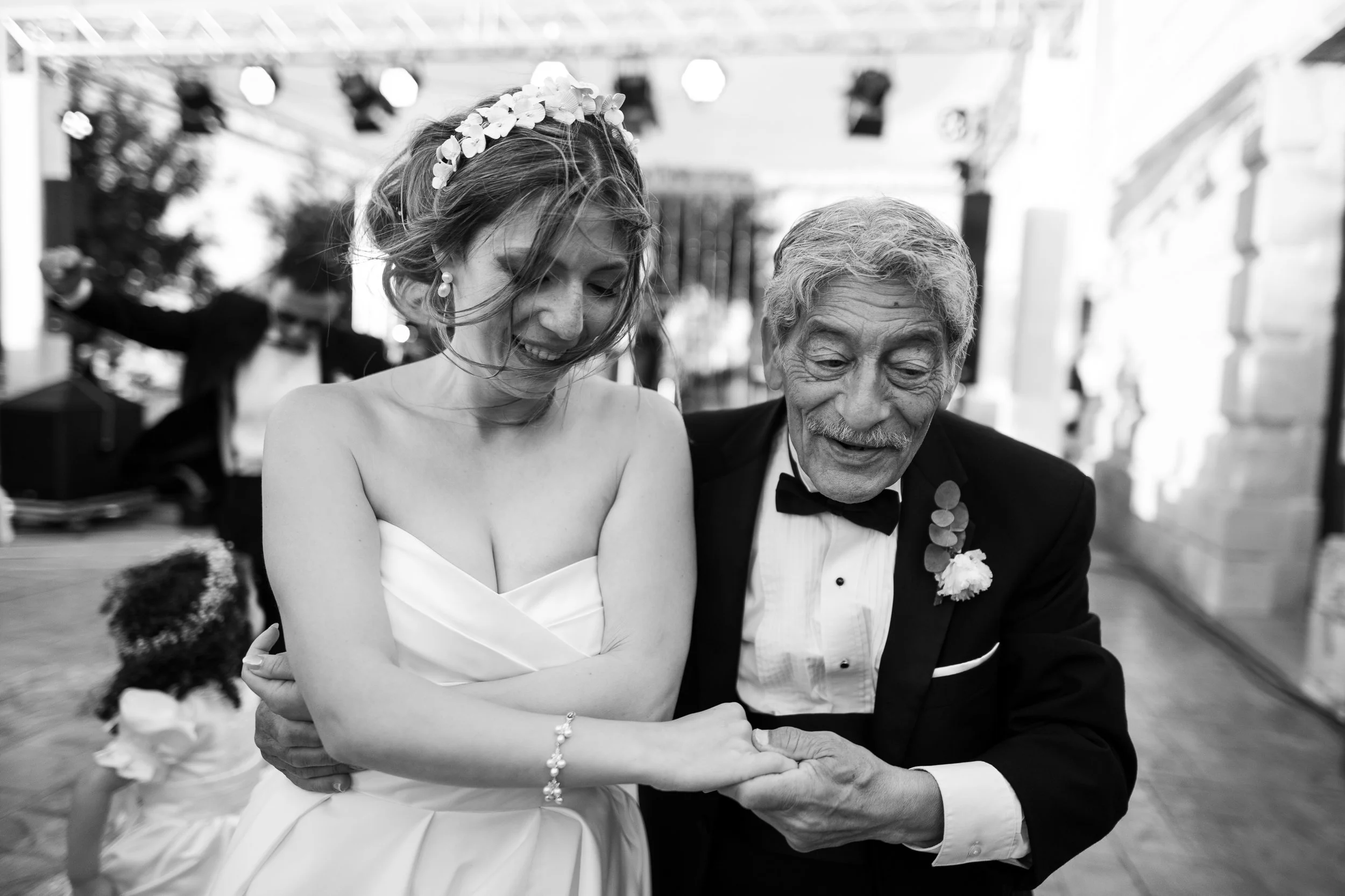 A bride and an elderly man, possibly her grandfather, share a dance at a wedding reception. The bride is smiling and wearing a strapless wedding dress and a flower crown, while the man is dressed in a tuxedo with a bow tie and boutonniere, smiling as