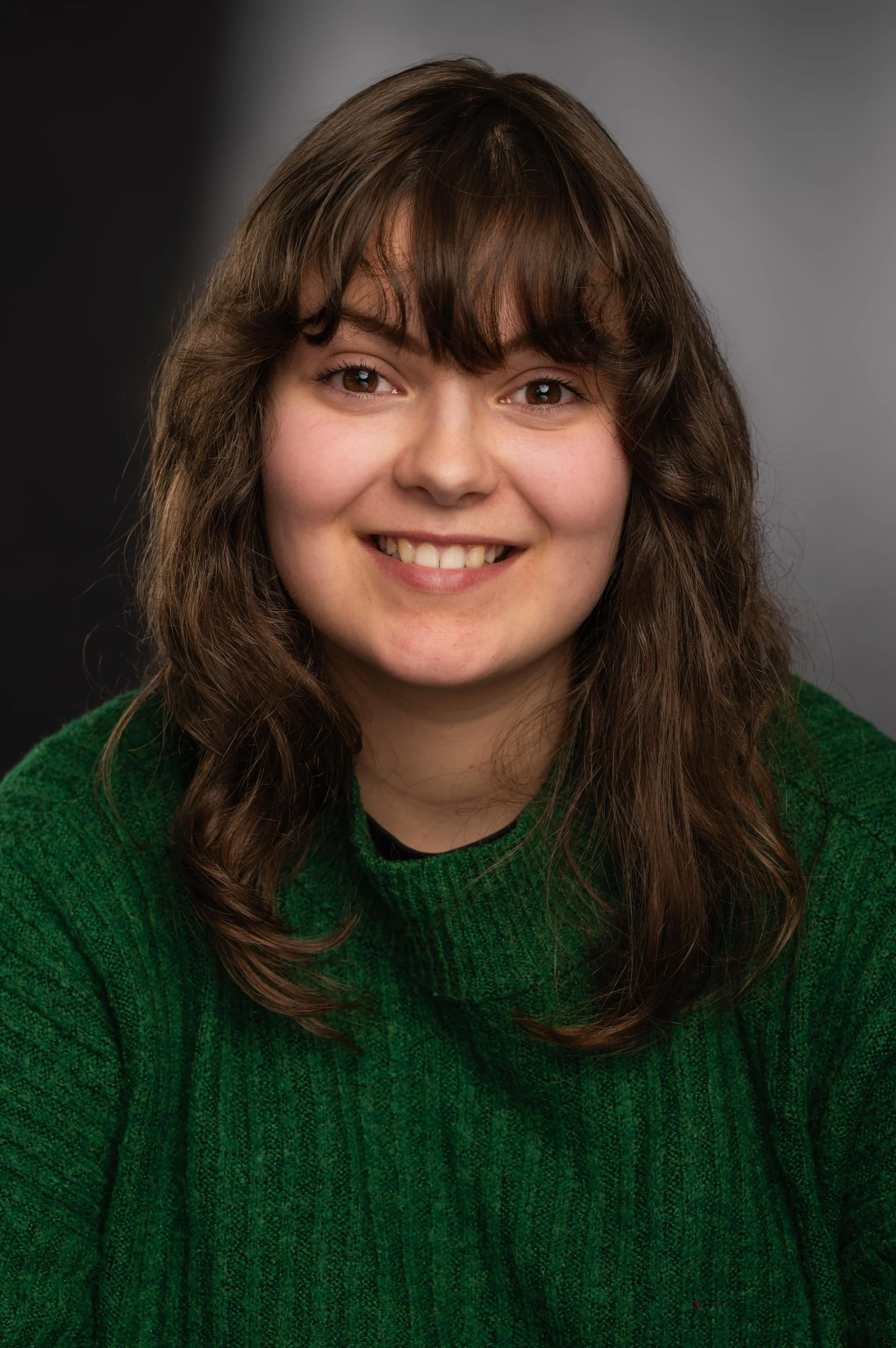A young woman with long brown curly hair, wearing a green sweater, smiling at the camera with a neutral-colored background.