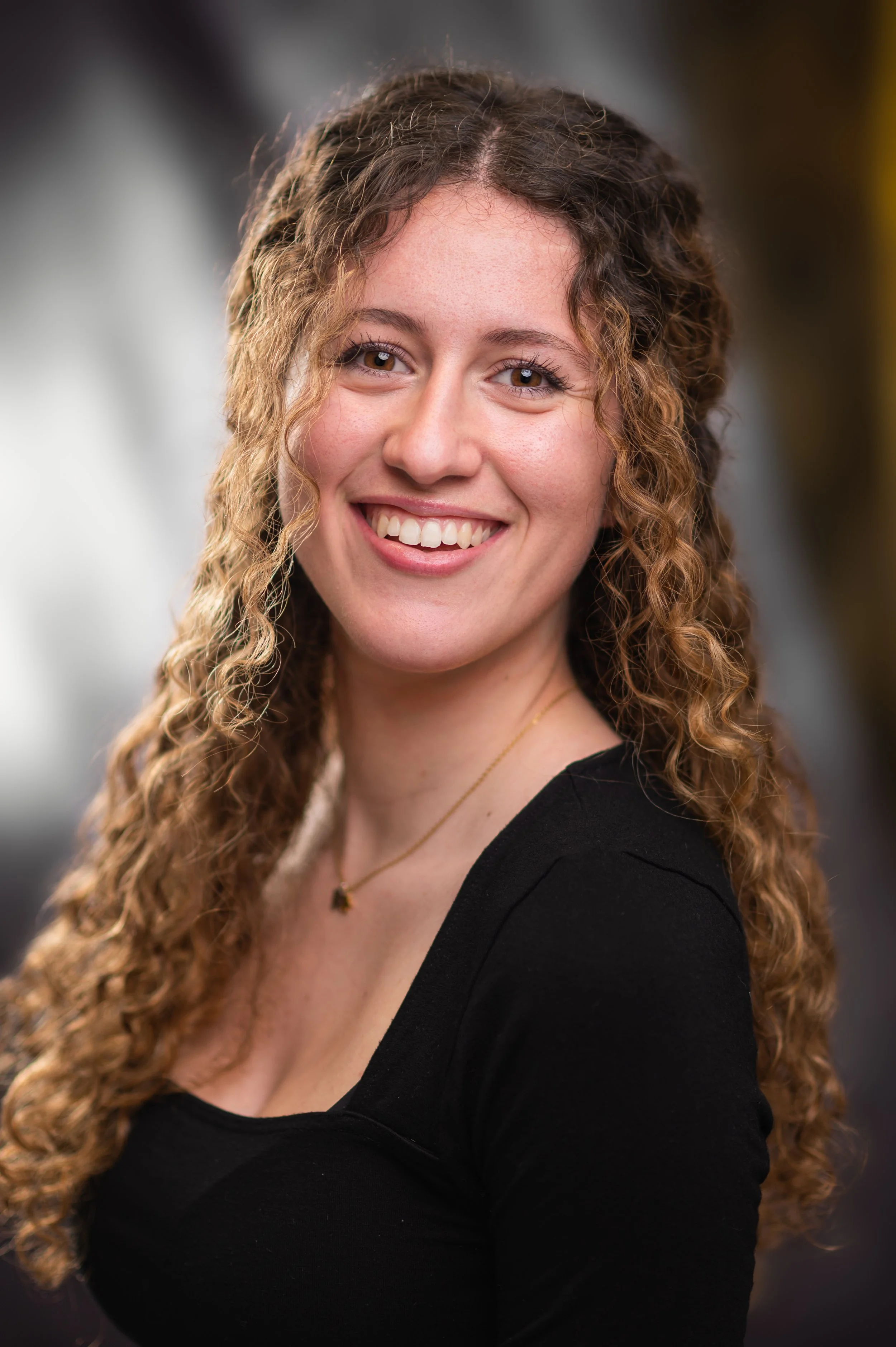 A woman with long, curly brown hair and hazel eyes, smiling and looking at the camera, wearing a black top and a delicate necklace.