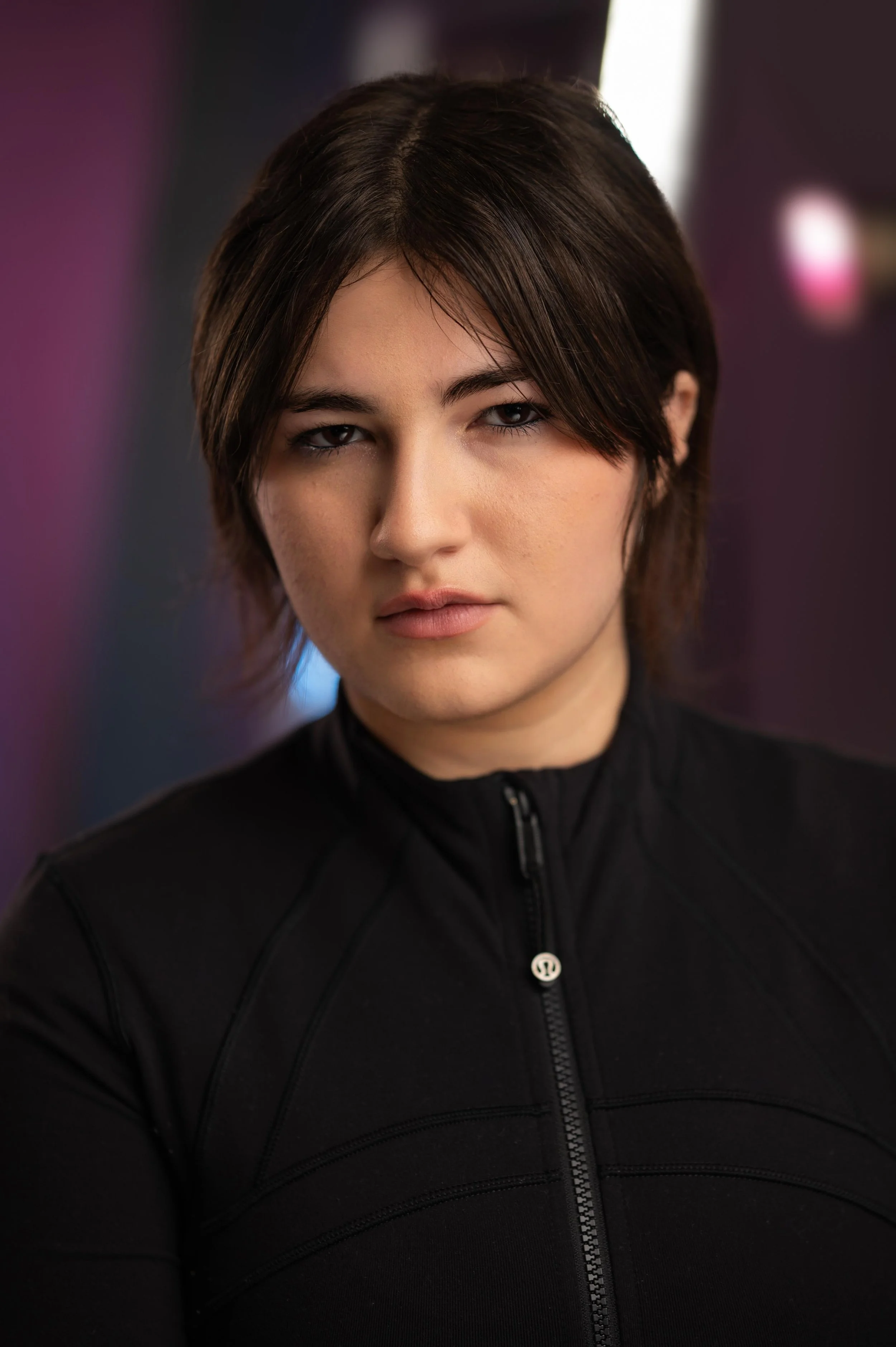 Close-up of a young woman with short, dark brown hair, wearing a black zip-up jacket with an Lululemon logo, looking directly at the camera with a serious expression, blurred purple and white background.