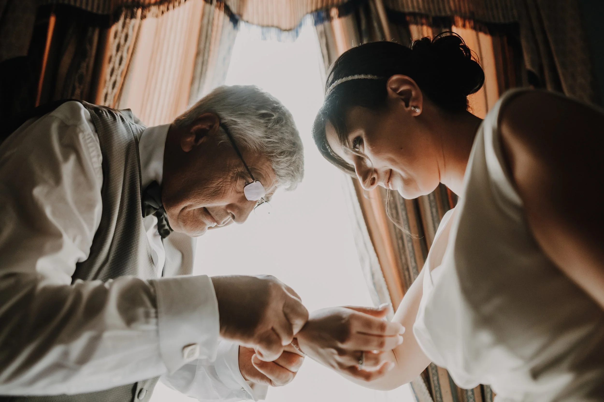 An elderly man and a young woman are leaning close together, holding hands, with their foreheads touching, sharing an intimate moment indoors with closed curtains and natural light coming through a window behind them.