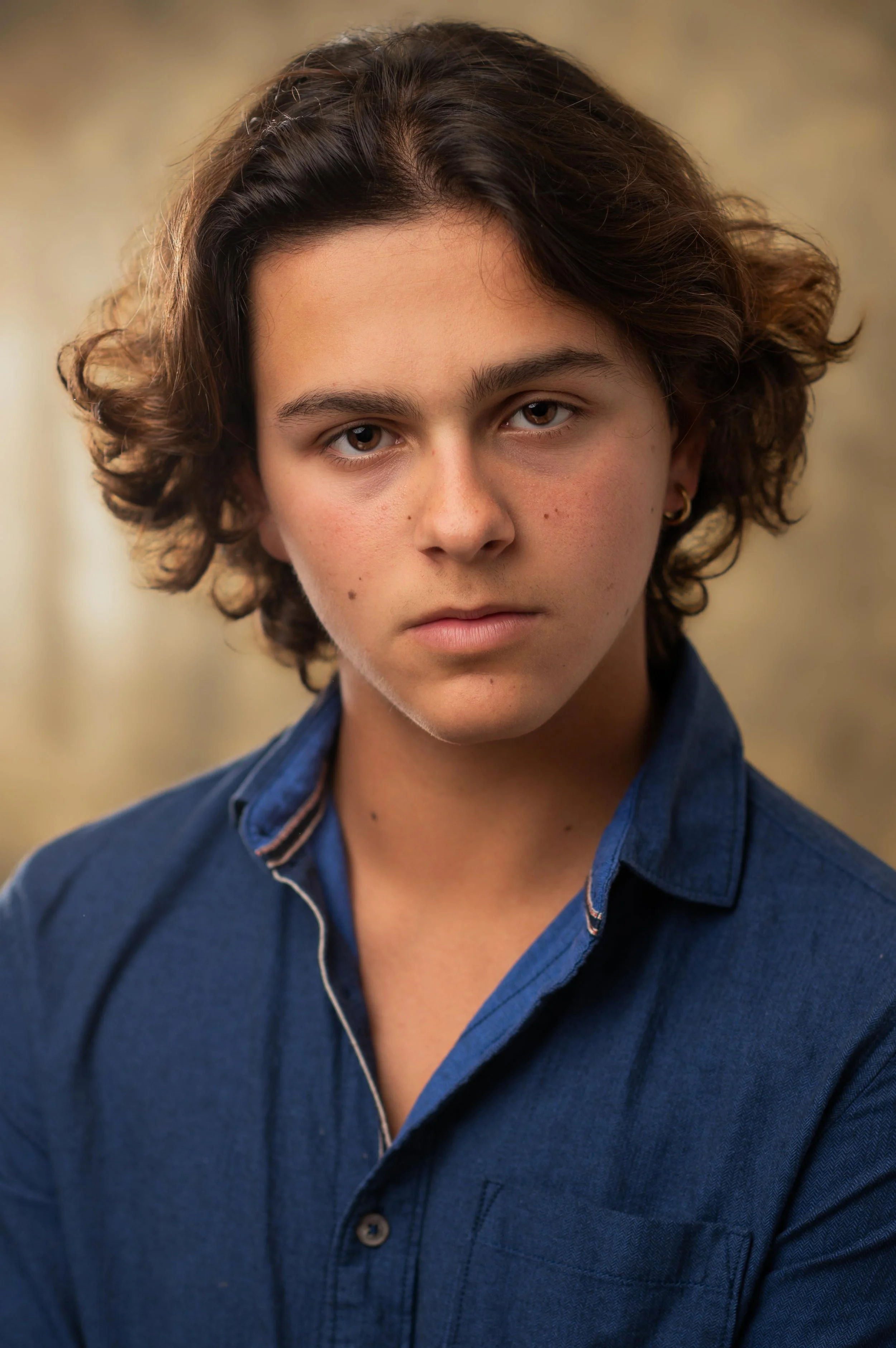 A young person with curly brown hair, wearing a dark blue shirt with a collar, looking directly at the camera with a neutral expression.
