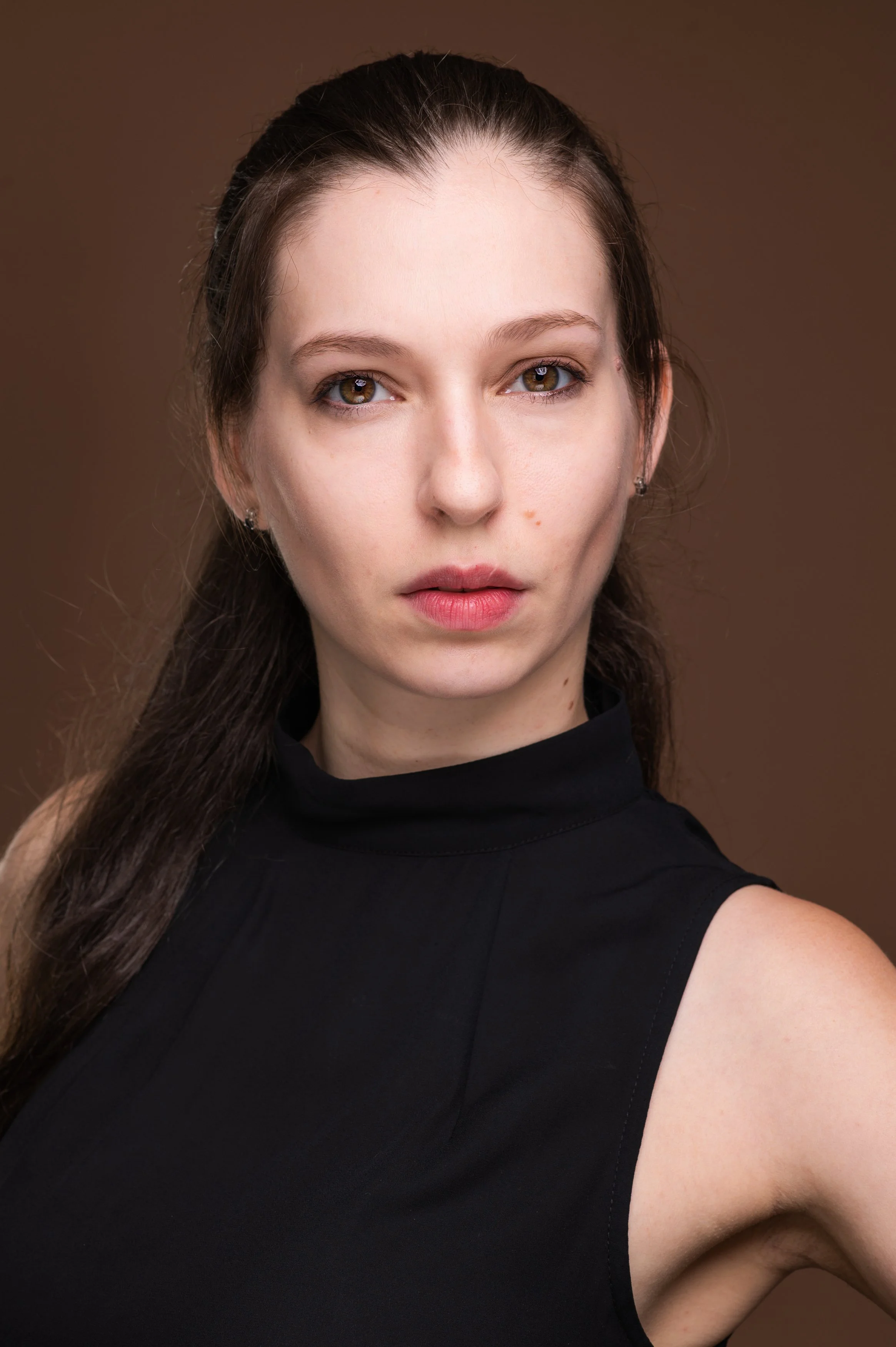 A young woman with long brown hair and light skin, wearing a black sleeveless top, looking directly at the camera against a brown background.