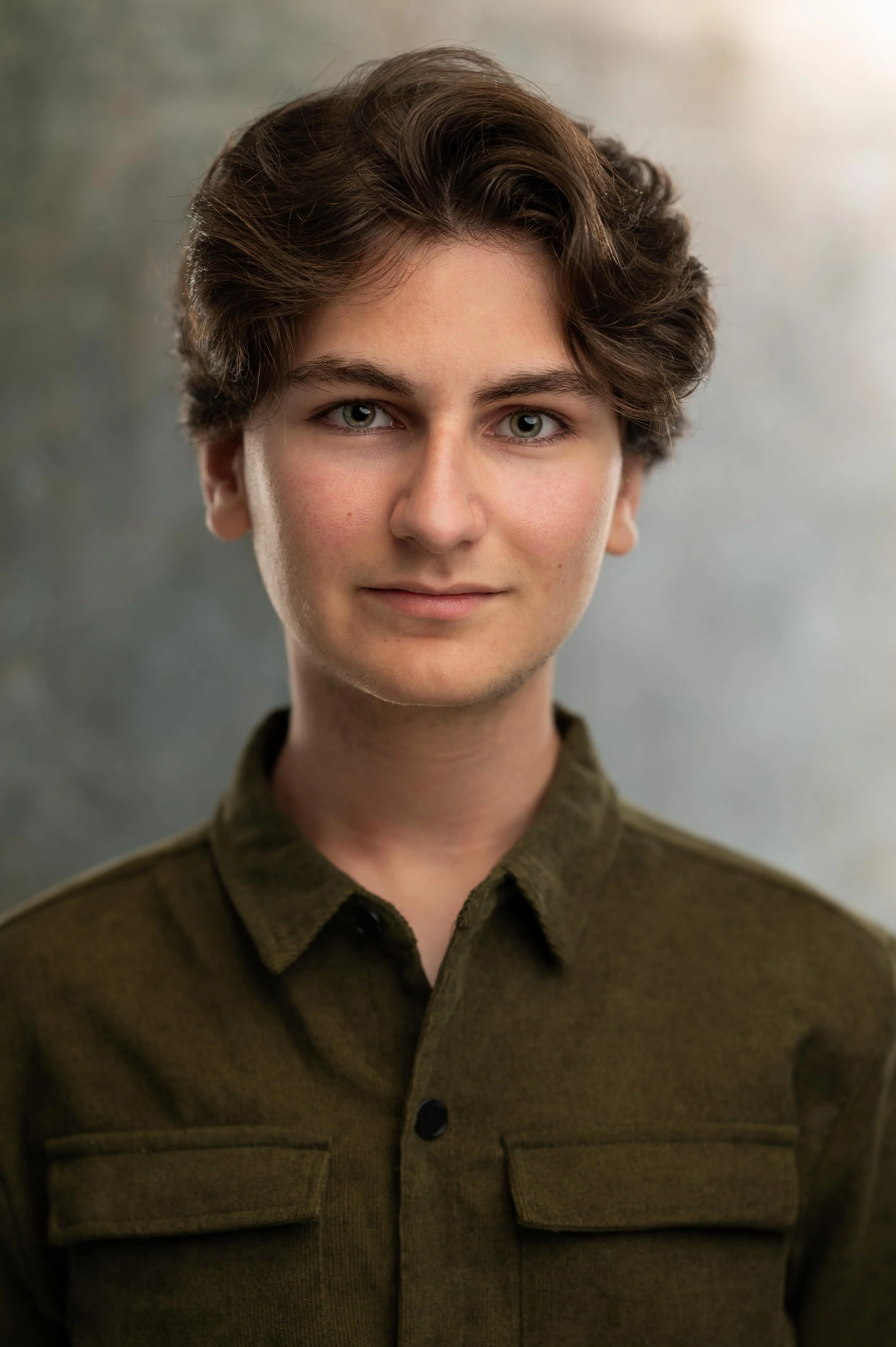 A young person with short brown curly hair, light skin, and green eyes wearing a dark green jacket with a collar and buttoned front, facing forward against a neutral background.
