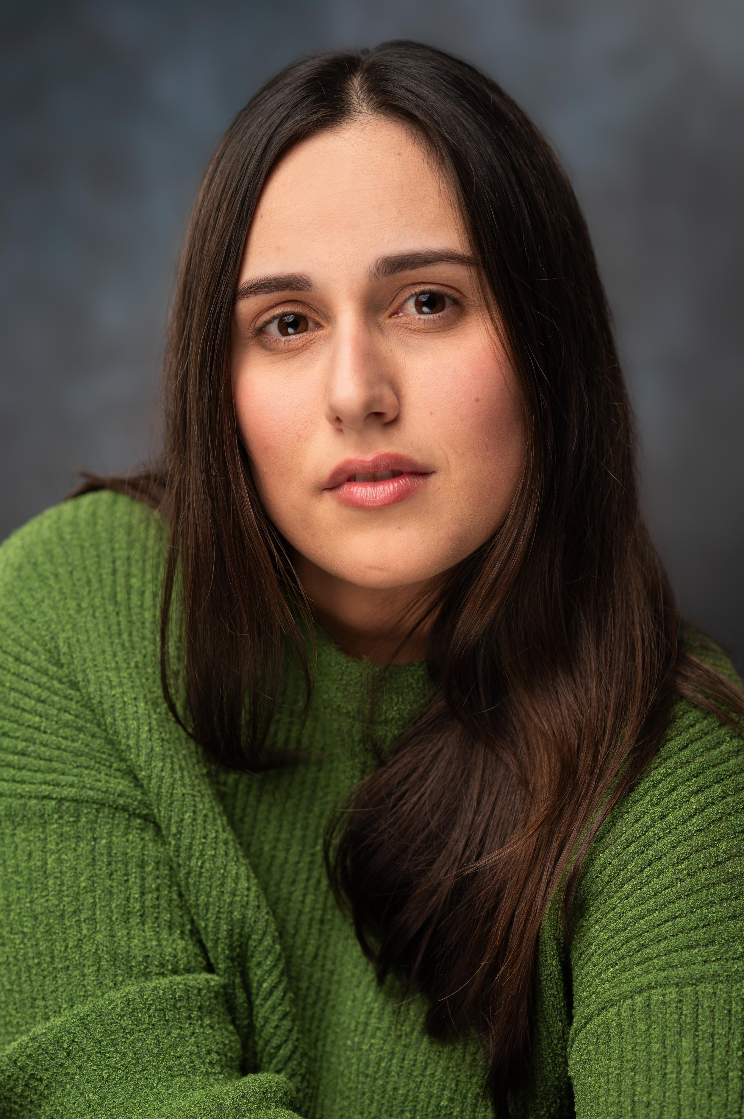 Close-up portrait of a young woman with long brown hair, wearing a green sweater, against a gray background.