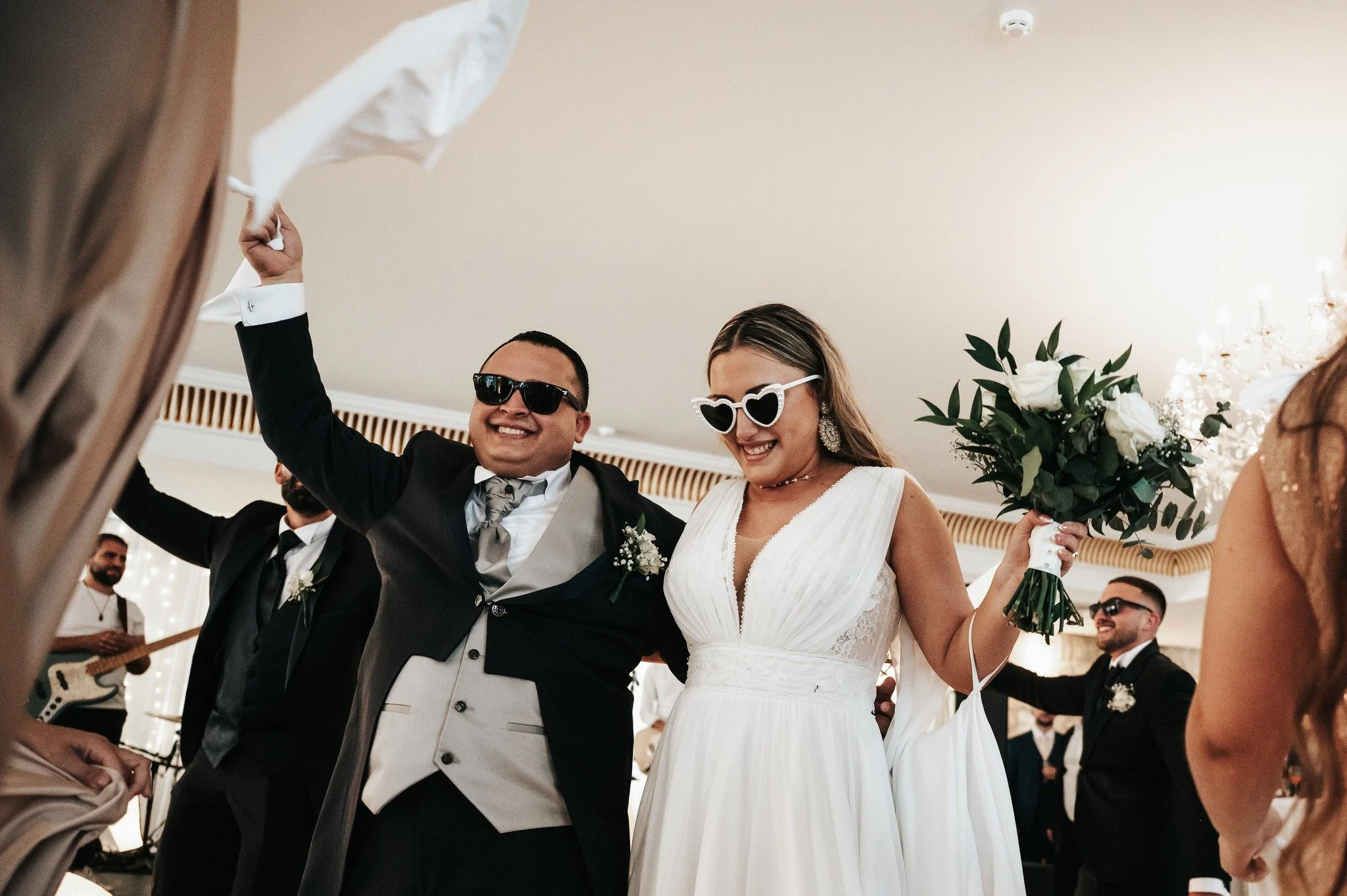 A bride and groom at their wedding reception smiling, wearing sunglasses, with the bride holding a bouquet of white flowers and green leaves, surrounded by guests in formal attire.
