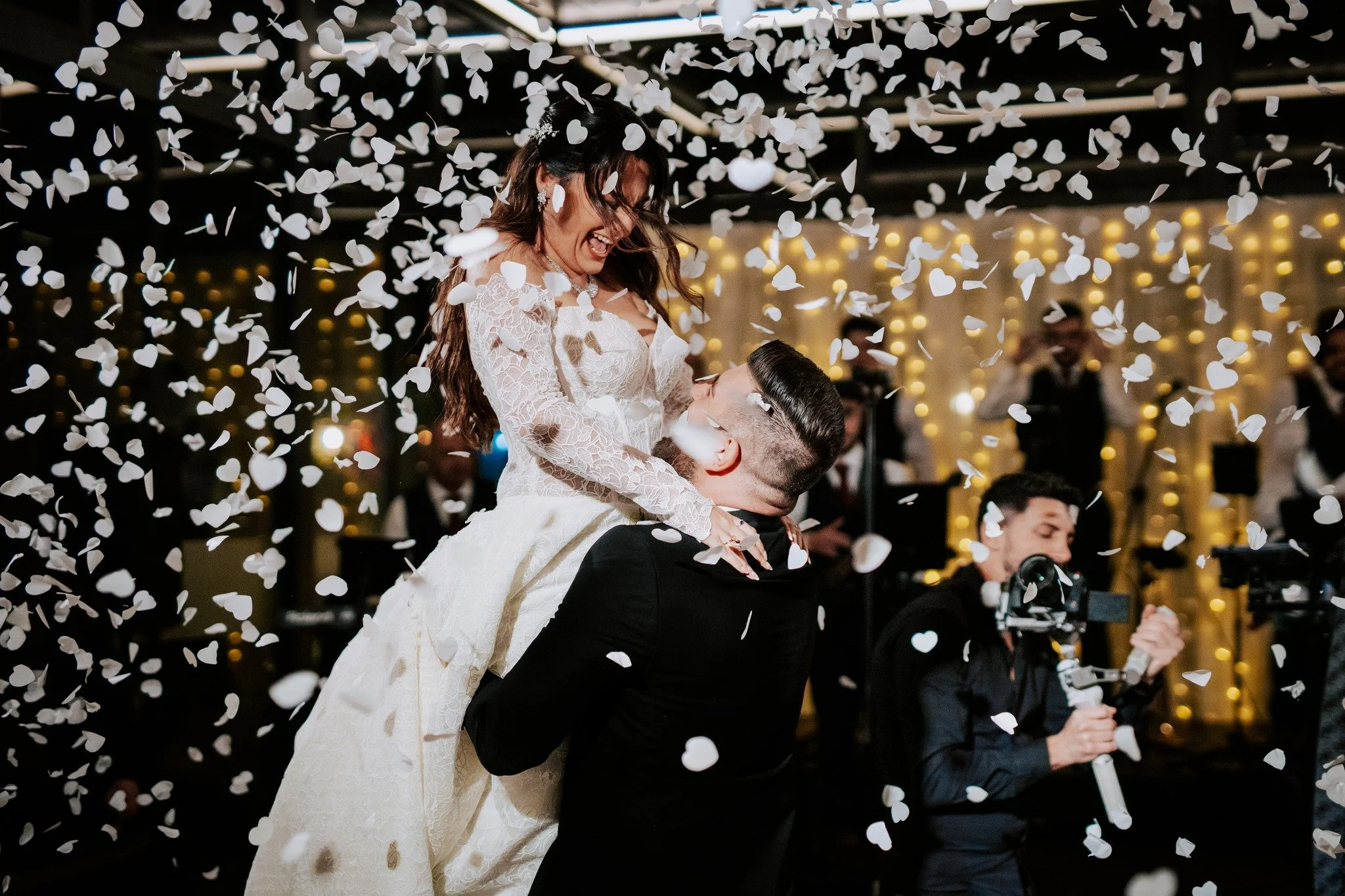 A bride and groom celebrating their wedding with confetti falling around them. The bride is smiling and wearing a white lace wedding dress, while the groom is lifting her up. In the background, other guests and wedding decorations are visible.