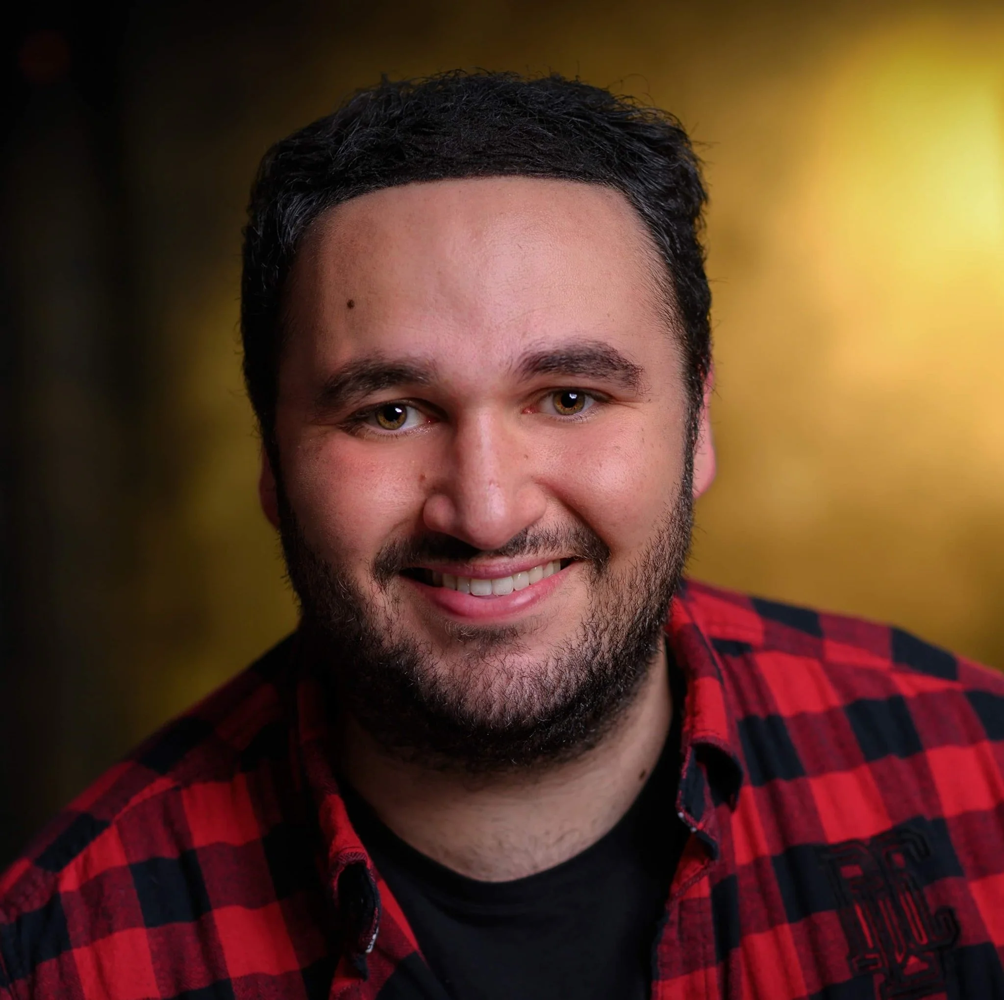 Close-up of a young man's face with light brown eyes, dark curly hair, and facial hair, smiling slightly, wearing a red and black checkered shirt, against a blurred yellow background.