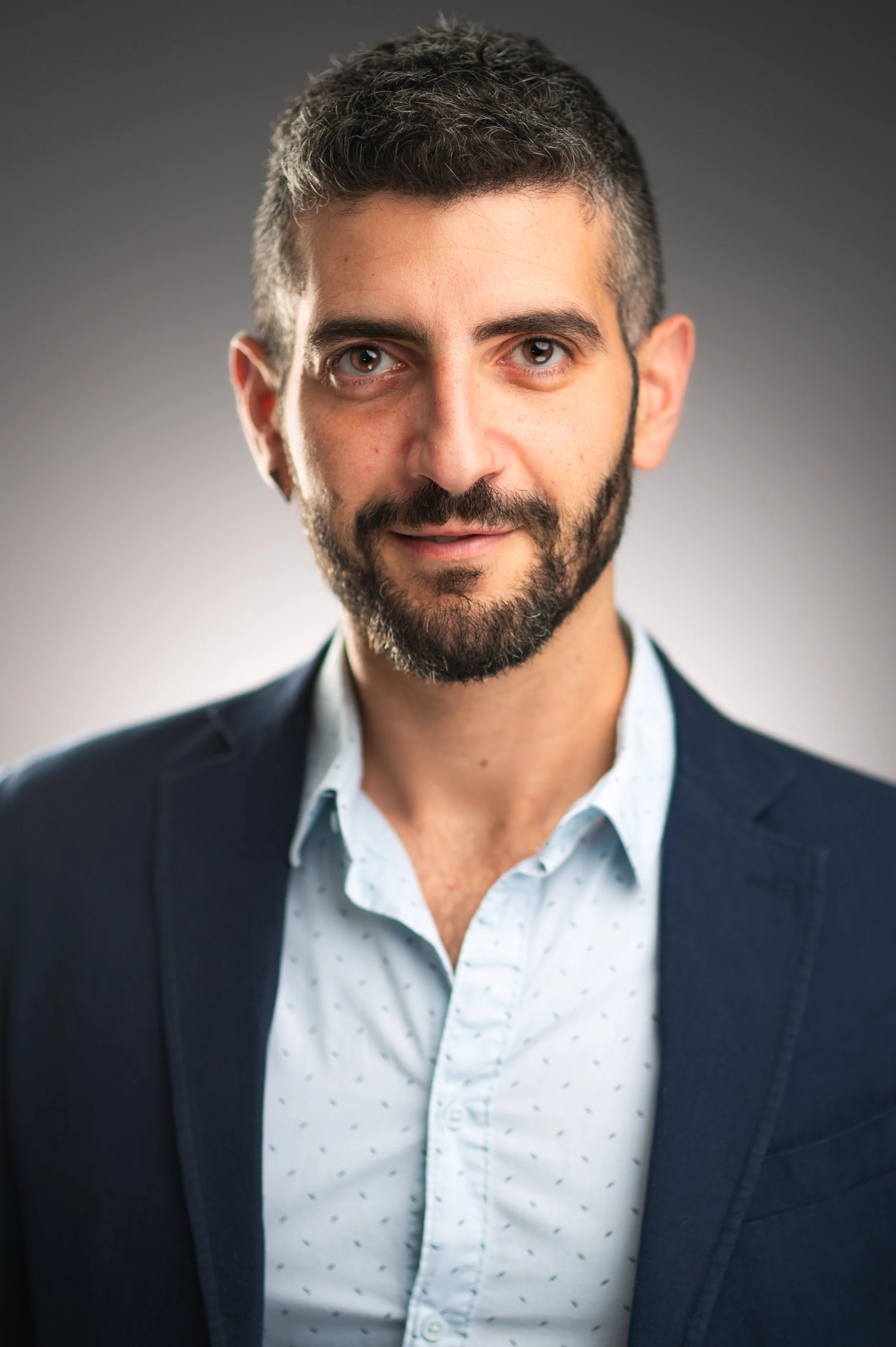 Close-up of a man with dark hair and a beard wearing a navy blazer and a white shirt with small dark patterns, against a gray gradient background.