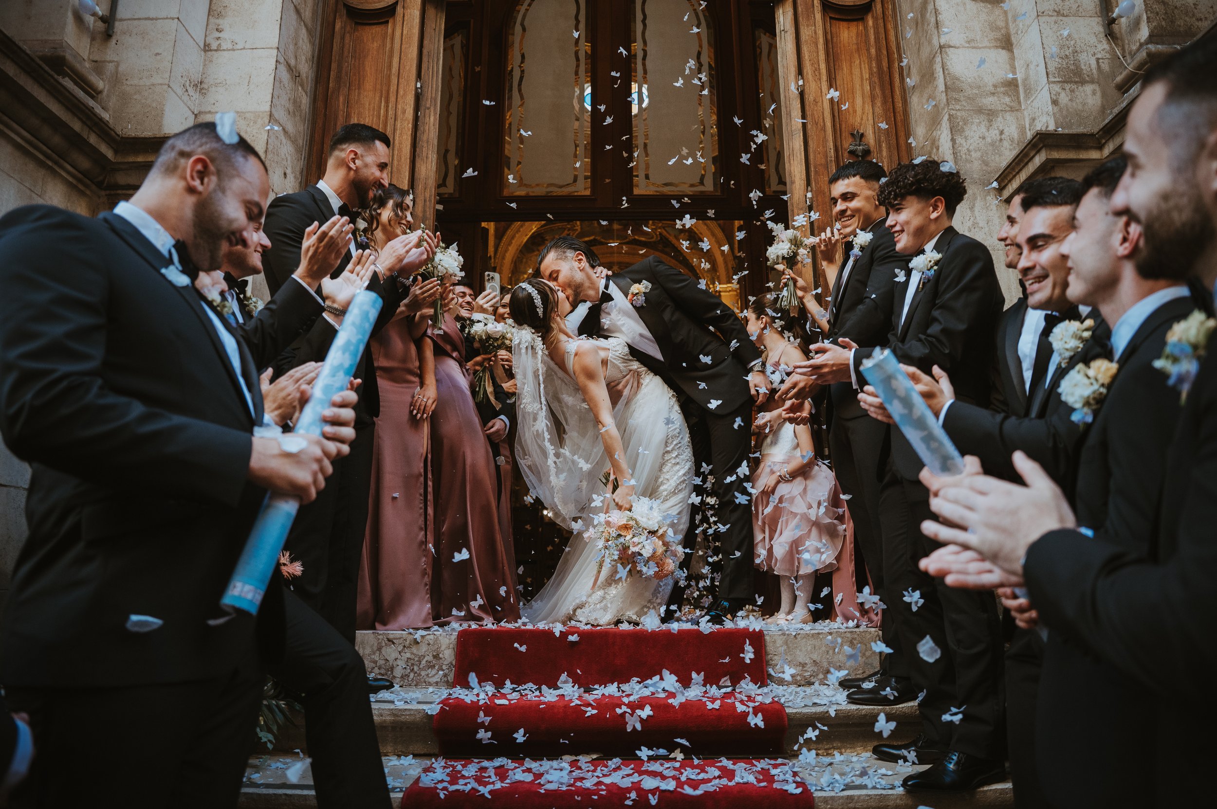 A bride and groom kiss on the steps of a historic building during their wedding celebration as friends and family celebrate around them, with white confetti falling and others holding bouquets and celebration items.