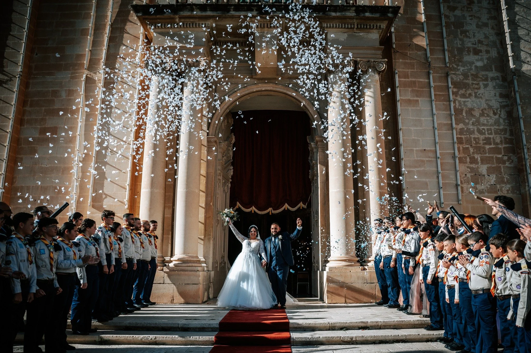 A newlywed couple stands on a red carpet outside a stone building, celebrating their wedding with guests on either side throwing white flower petals. The bride is wearing a white wedding dress and the groom is in a dark suit. The crowd is clapping and smiling.