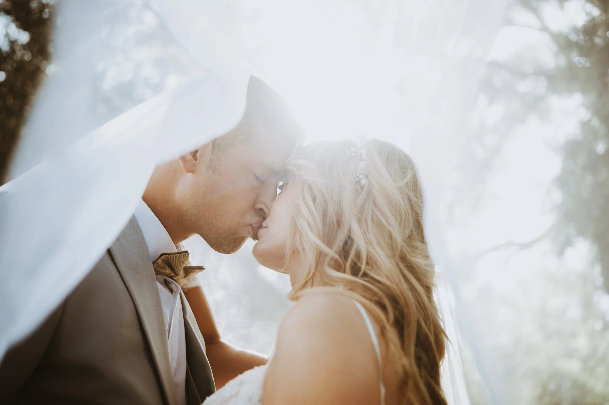 A couple shares a kiss outdoors, with the man in a gray suit and bow tie and the woman in a white dress, surrounded by trees and sunlight.