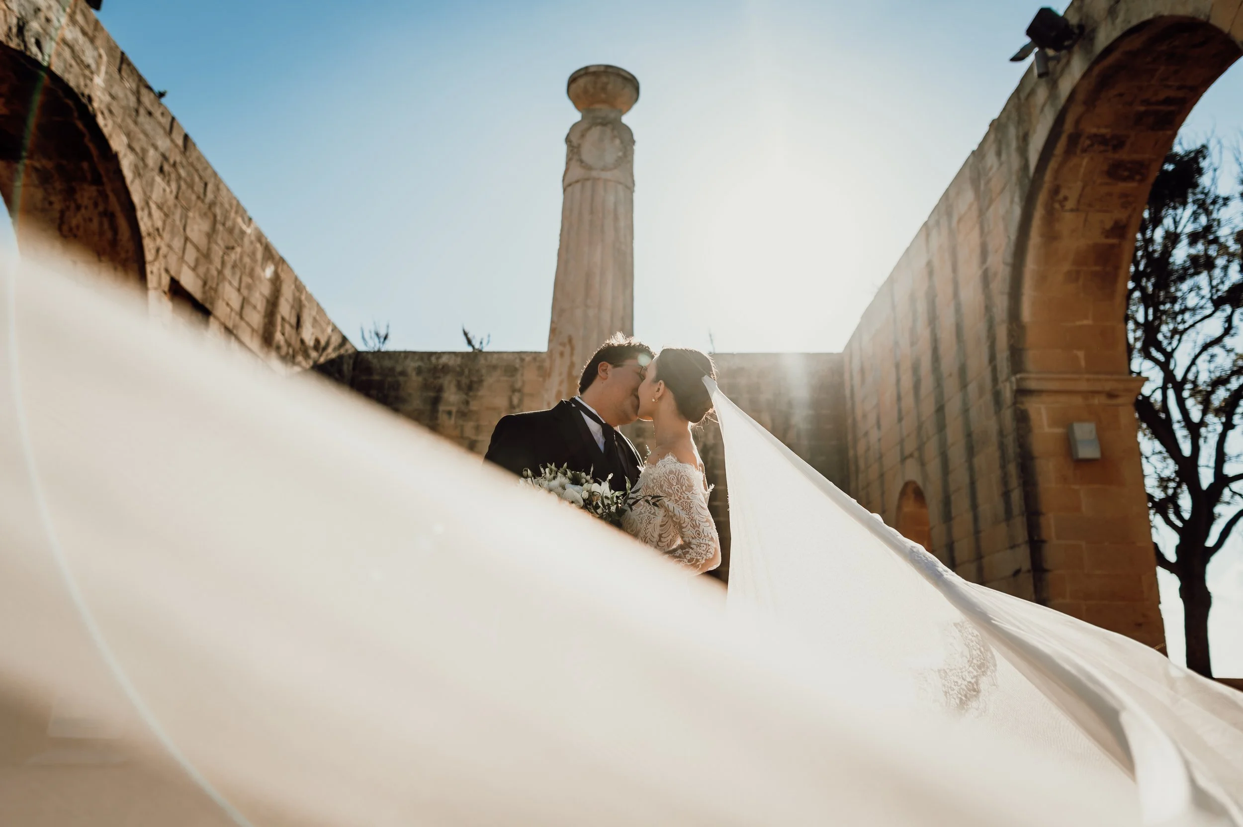 A bride and groom sharing a kiss during their wedding in an outdoor historical site with stone arches and a tall stone pillar.