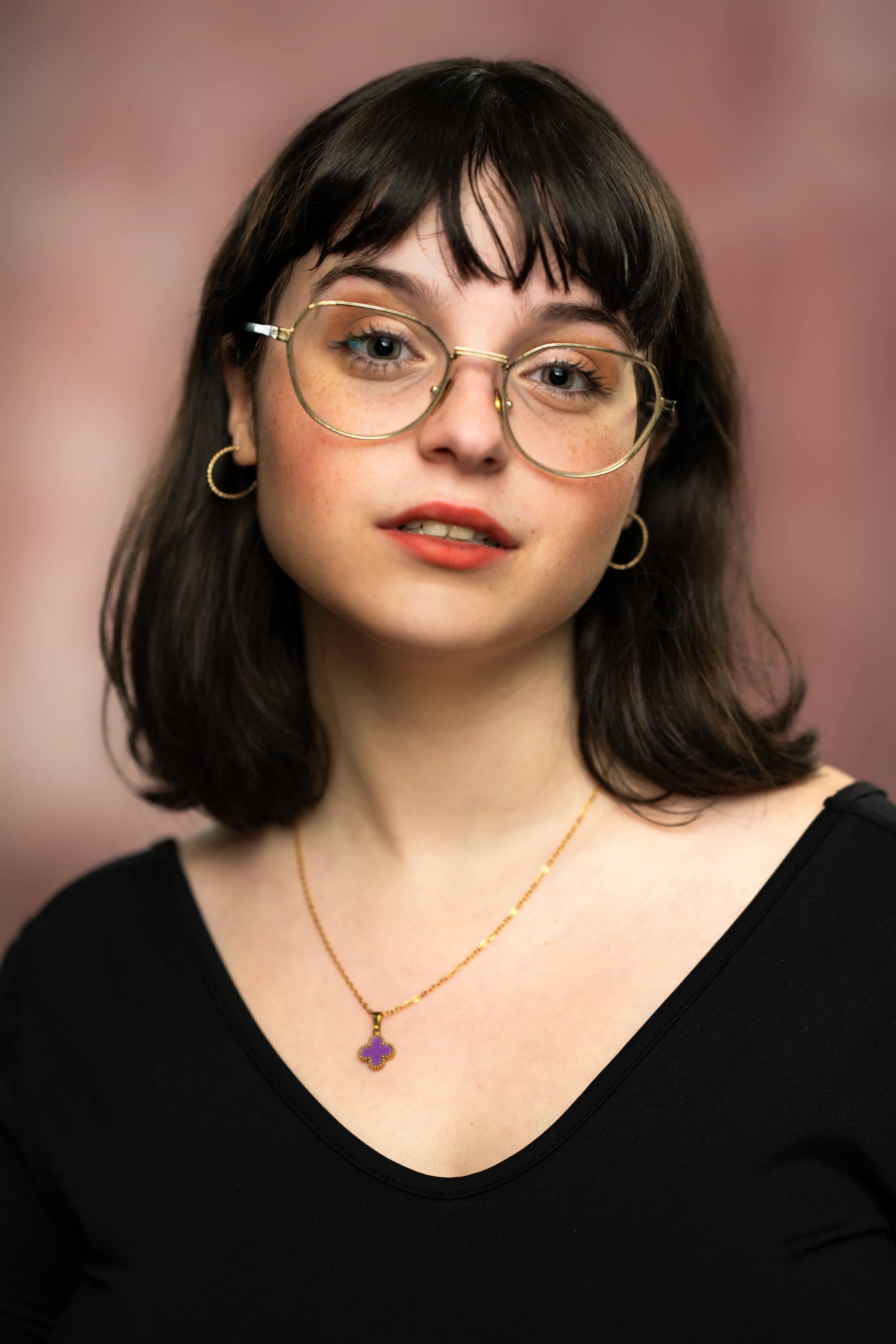 Close-up of a young woman with dark hair, wearing glasses, gold hoop earrings, a gold necklace with a four-leaf clover pendant, and a black top, against a pink background.