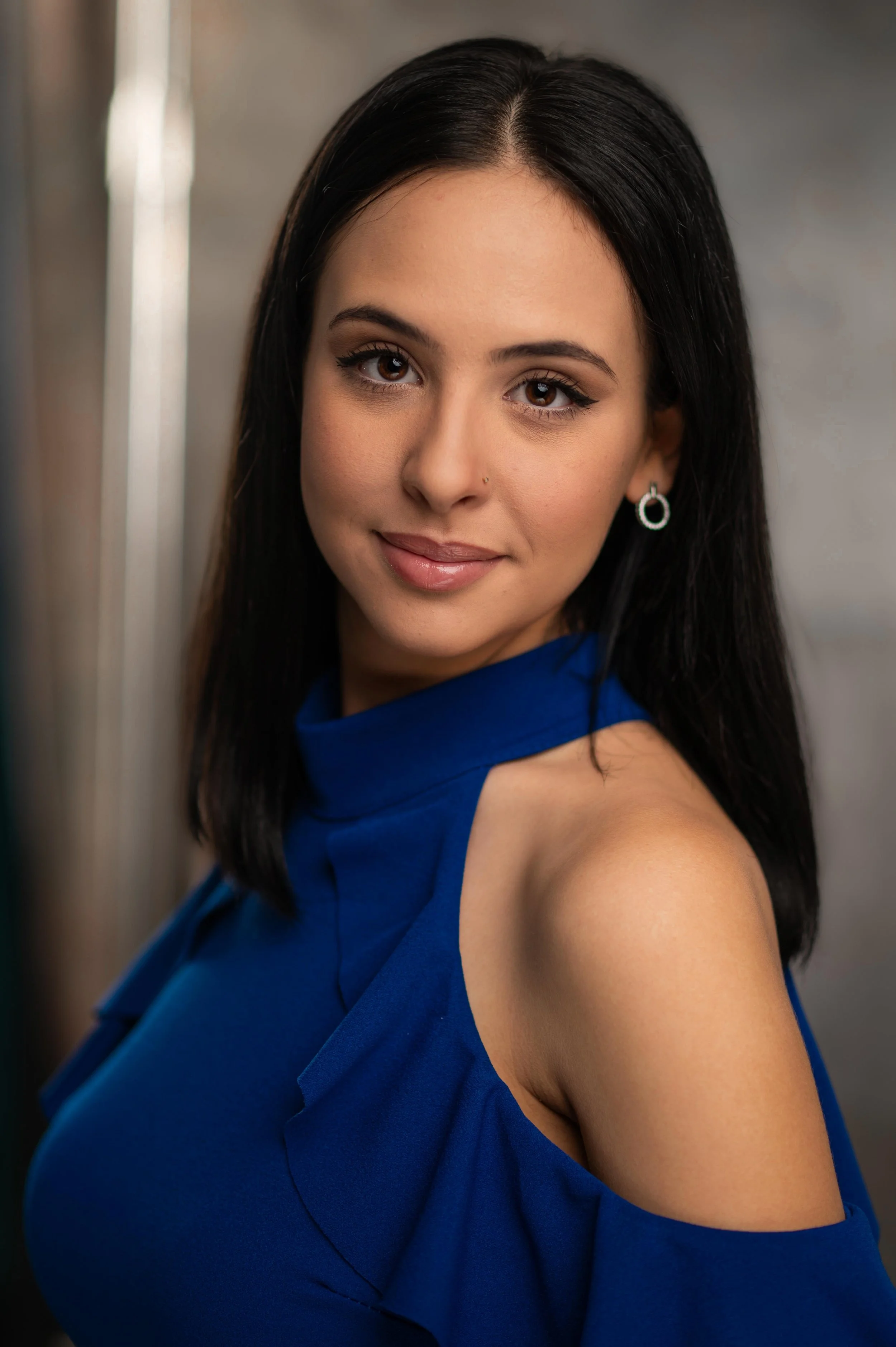 A woman with long black hair, brown eyes, wearing a blue off-the-shoulder dress and earrings; smiling softly with makeup, in a portrait shot.