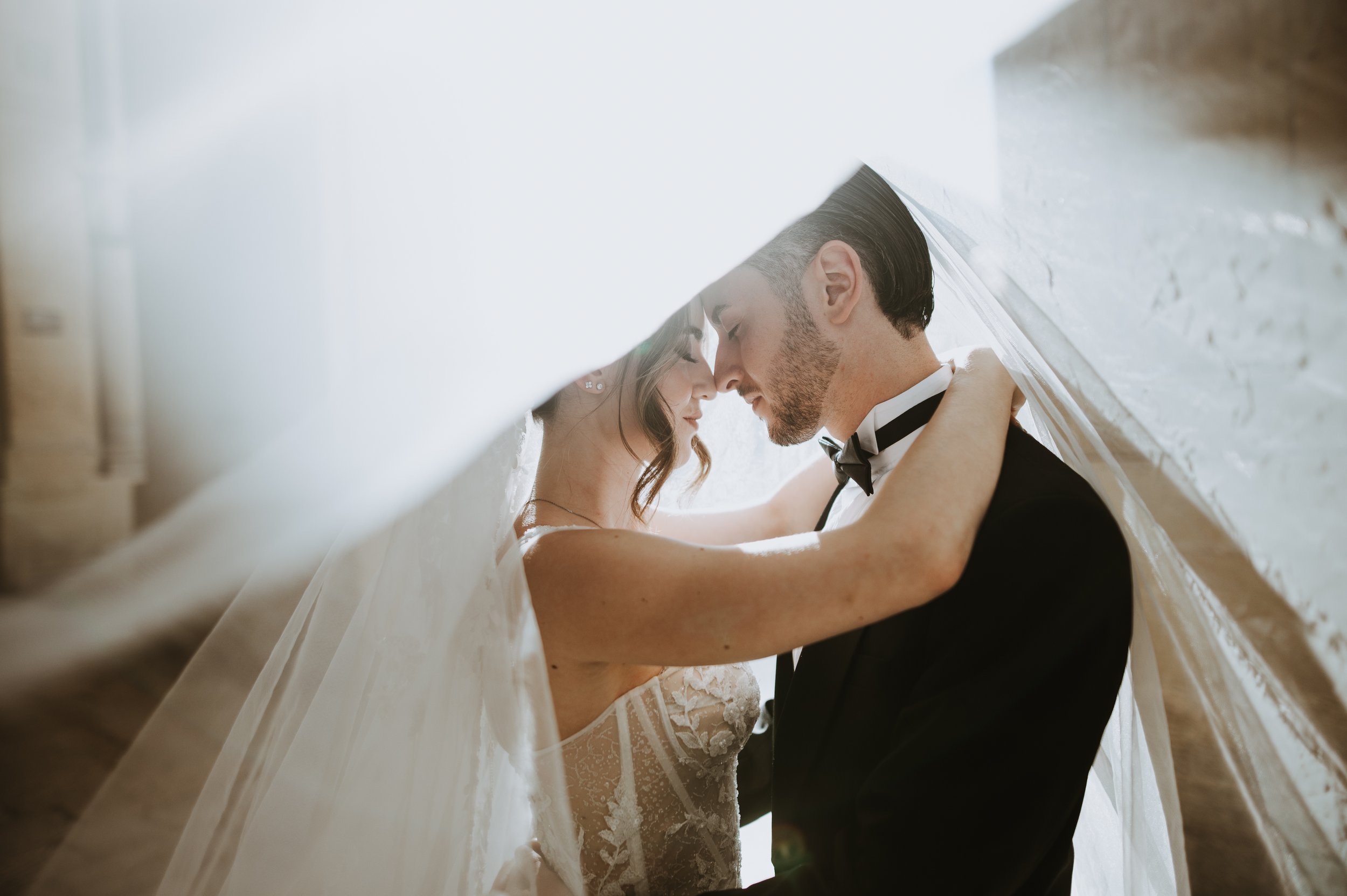 A bride and groom are kissing under a wedding veil, with the bride in a lace wedding dress and jewelry, and the groom in a tuxedo and bow tie, standing indoors by a white brick wall.