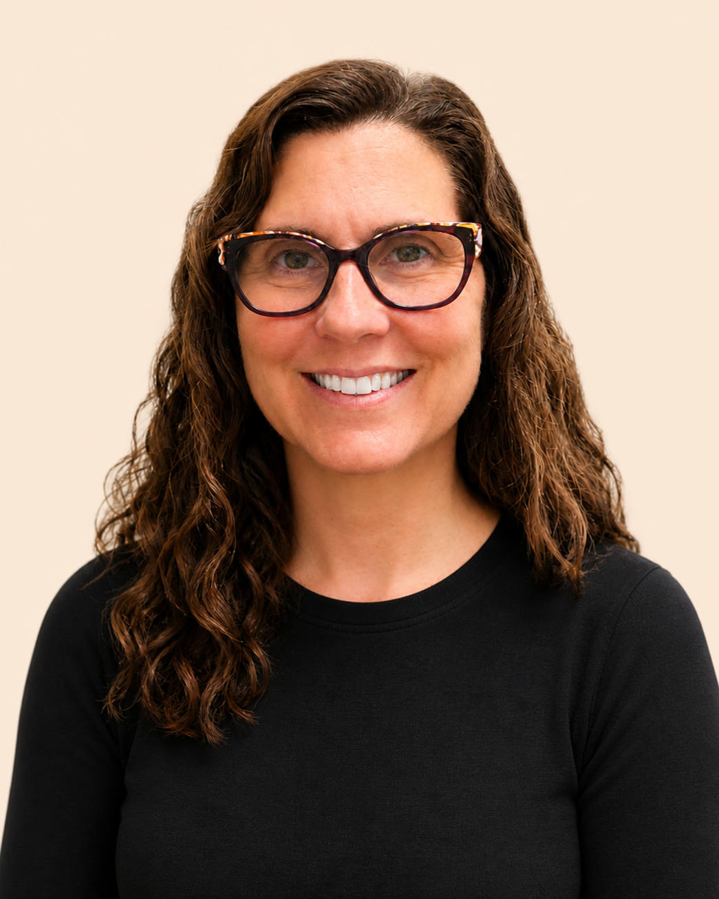 A woman with curly brown hair, wearing black-rimmed glasses, smiling, dressed in a black top, standing against a plain light-colored background.