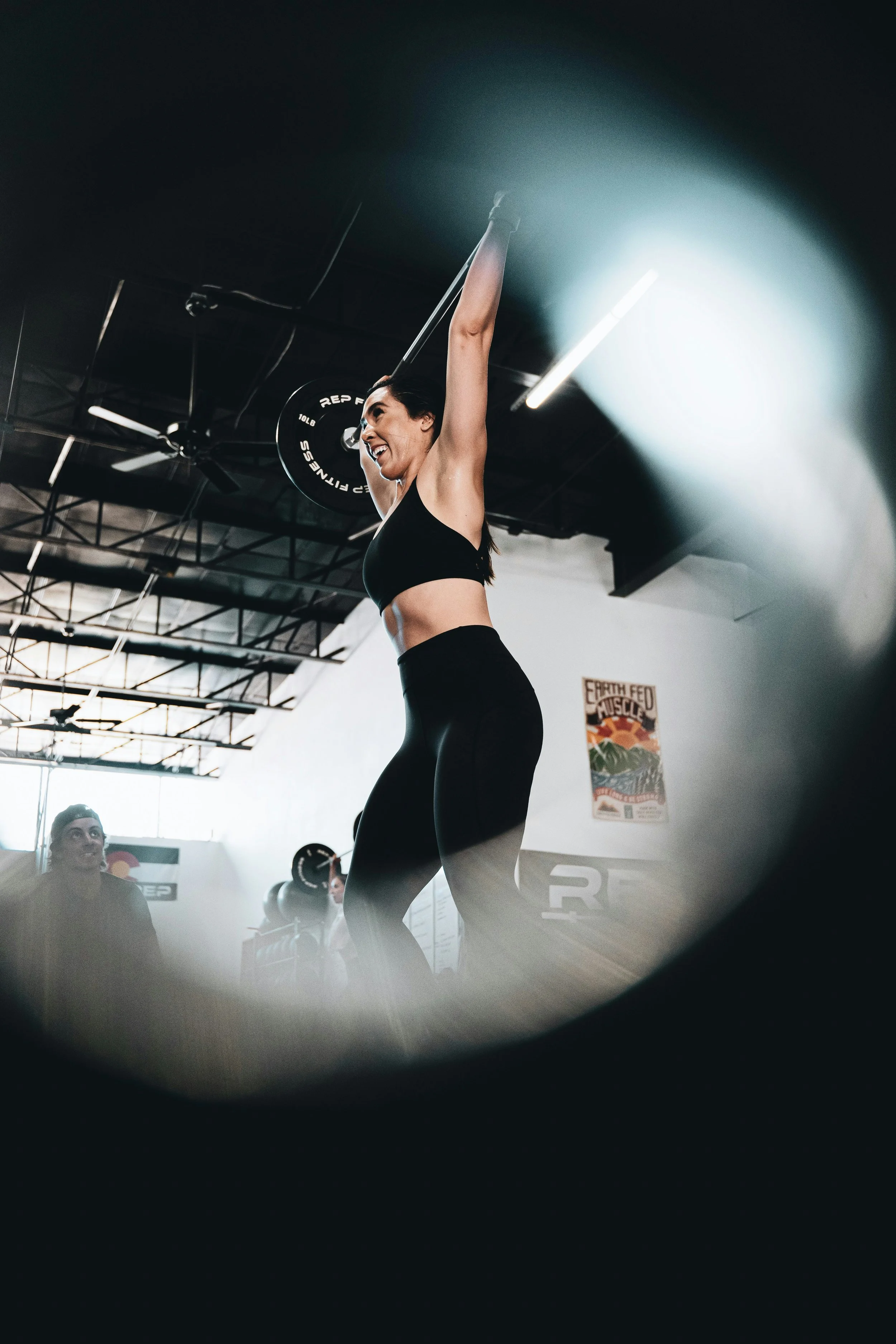 A woman in athletic wear lifting a barbell overhead in a gym with industrial ceiling and fluorescent lighting, viewed through a circular frame.