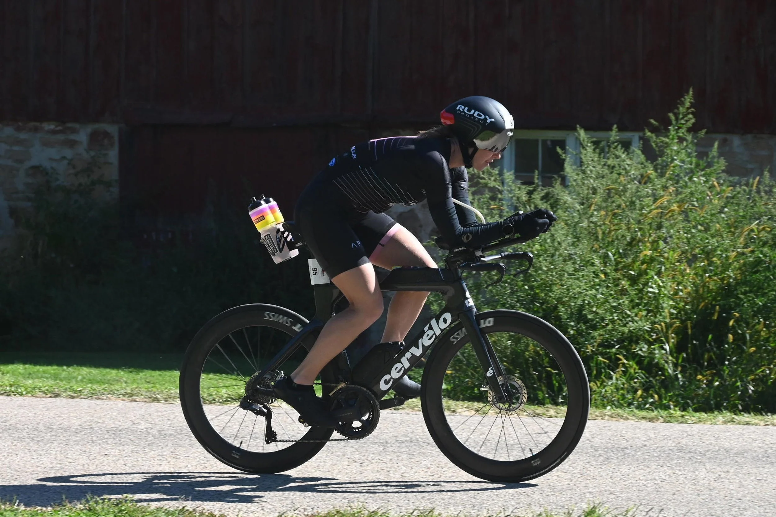 Person riding a black Cervélo road bike on a paved path, wearing a black helmet with 'Rudy' written on it, black cycling gear, and sunglasses, with greenery and a house in the background.