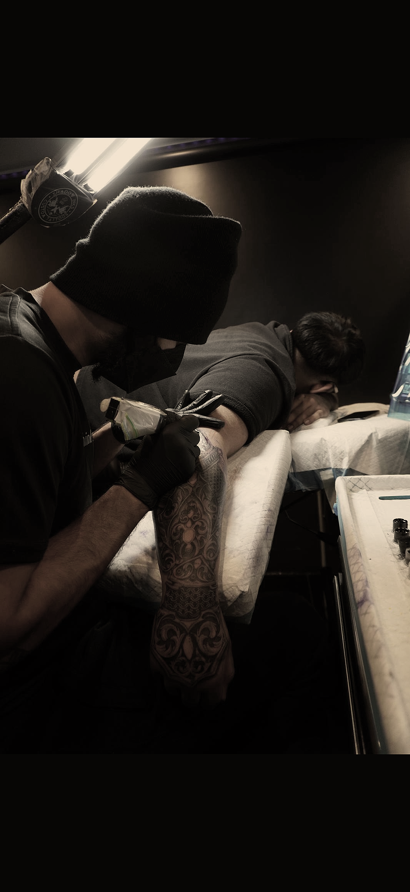 A person receiving a tattoo on their arm by a tattoo artist in a dimly lit environment.