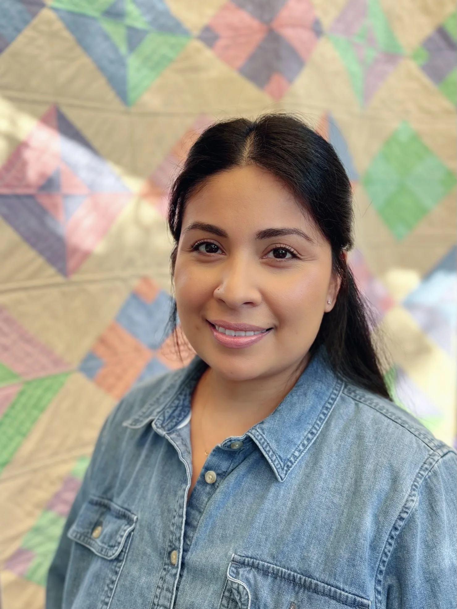 Picture of a young woman with dark hair wearing a denim jacket, smiling in front of a colorful, geometric-patterned quilt.