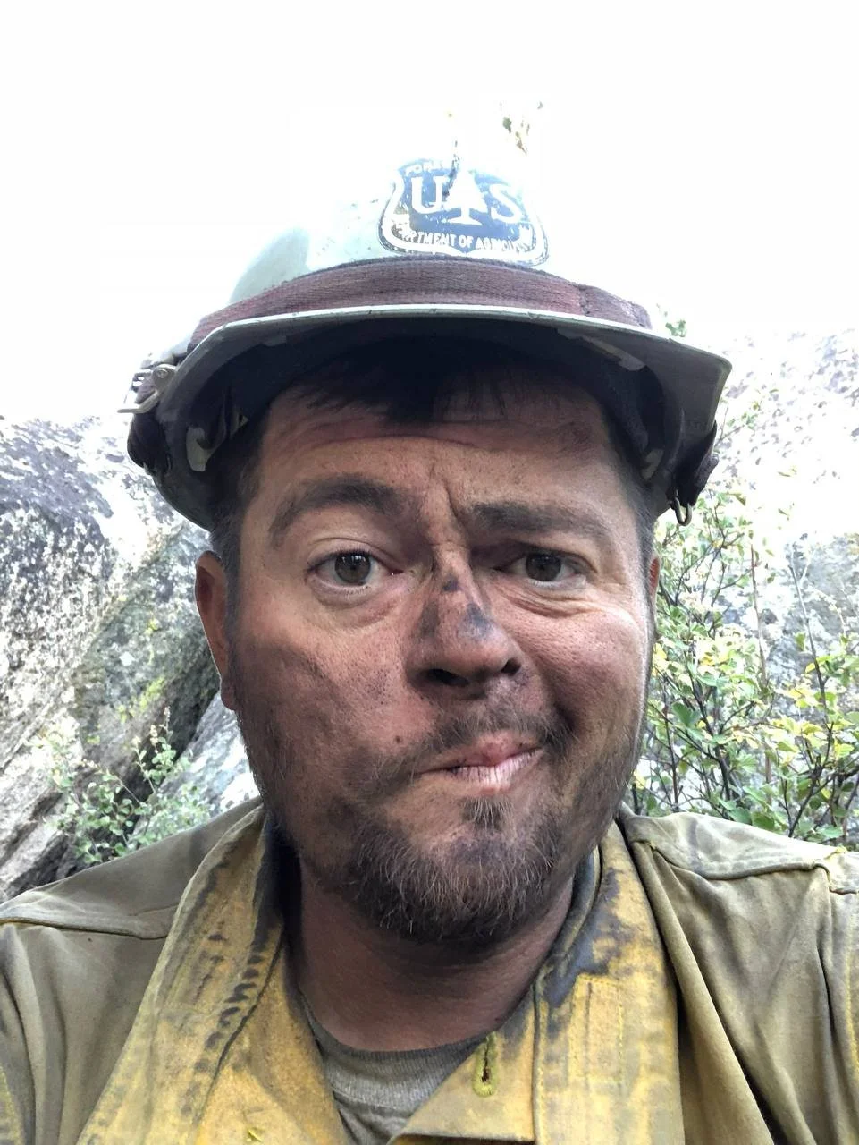 A firefighter with soot-covered face and uniform, wearing a helmet with the US Forest Service emblem, outdoors in a wooded area.