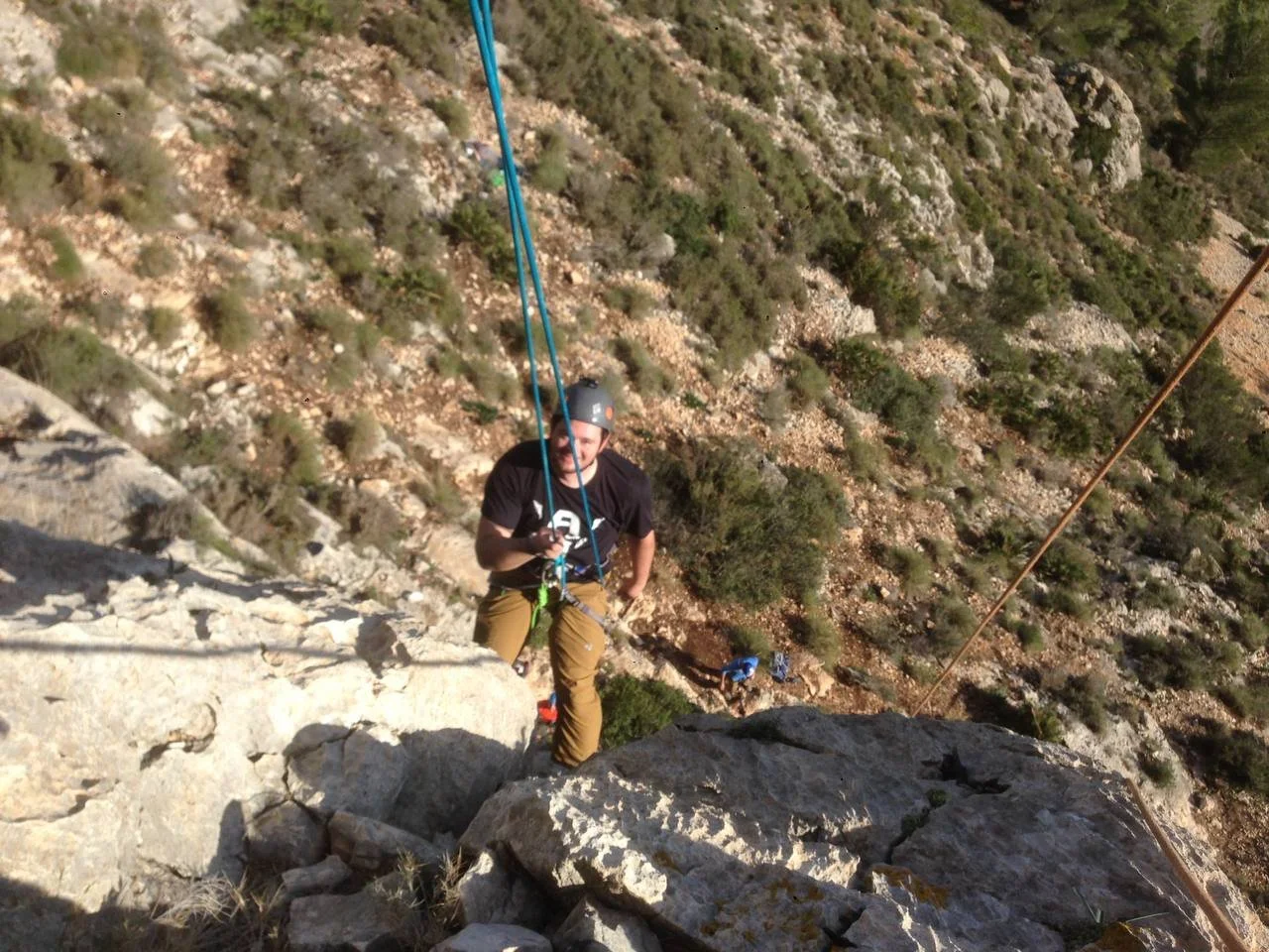 A person rock climbing on a steep outdoor cliff, wearing a helmet and safety harness, with a rocky and hilly landscape in the background.
