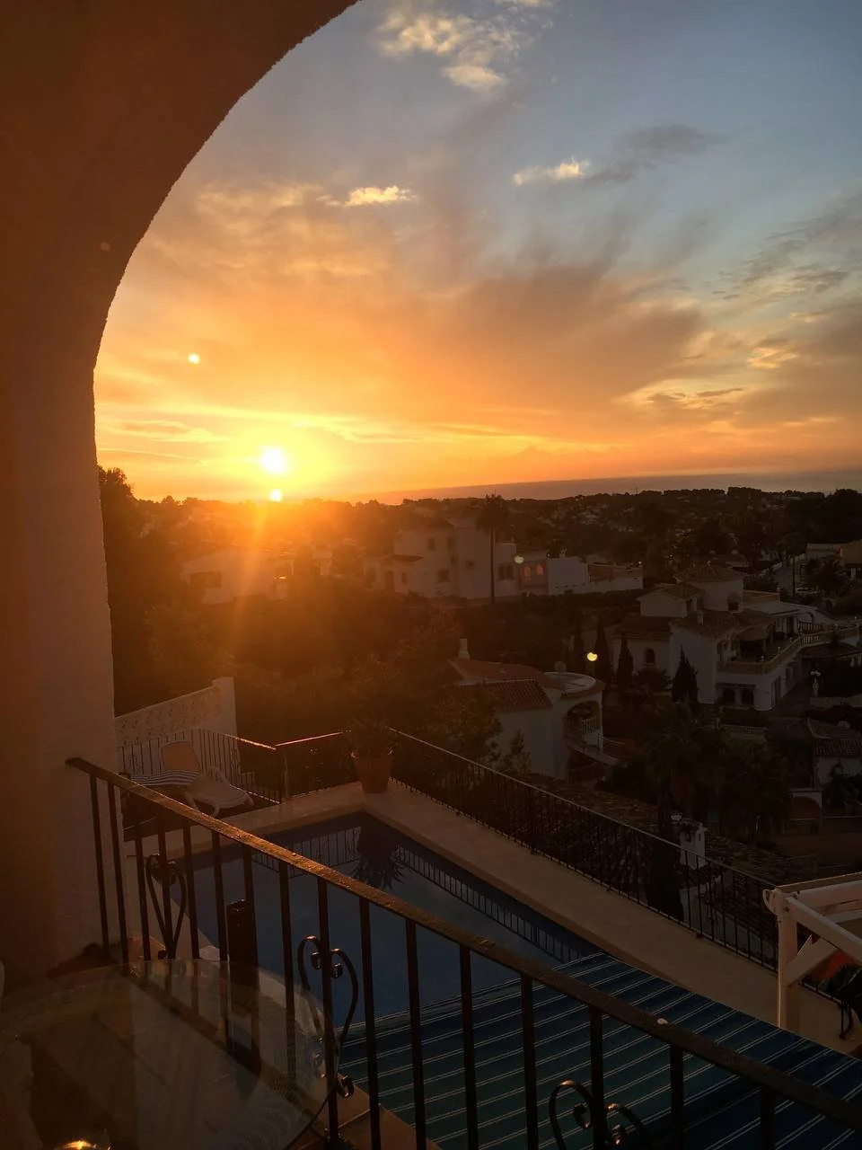 Sunset view from a balcony showing a pool, a potted plant, and a neighborhood with houses and trees against a colorful sky with clouds.