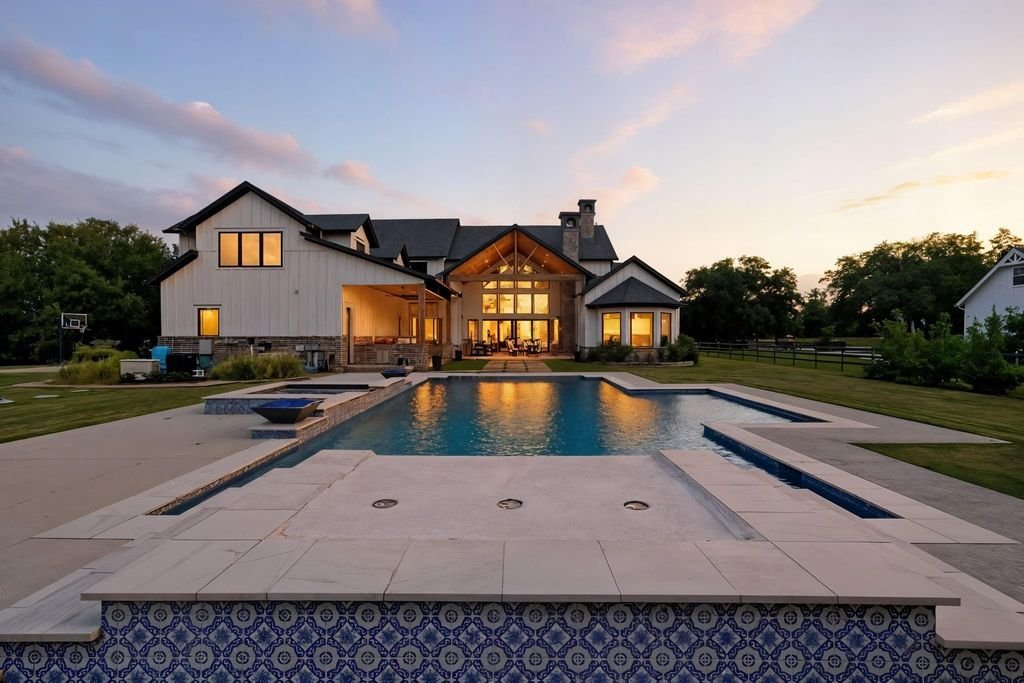 Large modern house with lit interior lights, outdoor pool in the front yard, green lawn, trees, and a sunset sky.
