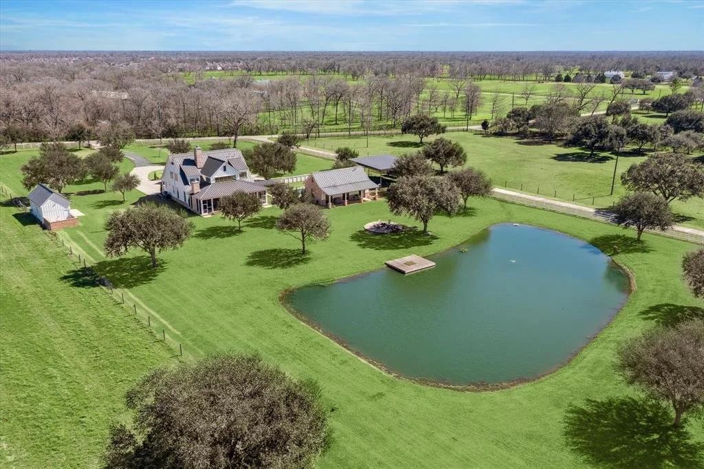 Aerial view of a large green backyard with a pond, trees, a house, a small shed, and separate garage, surrounded by open fields and distant trees under a partly cloudy sky.
