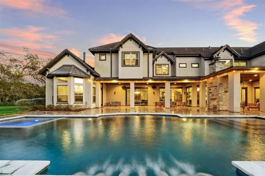 Large modern house with a backyard swimming pool at sunset, featuring a covered patio area with seating and large windows.
