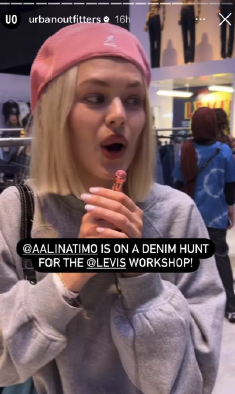 A young woman with blonde hair wearing a pink baseball cap and a grey sweatshirt, looking surprised or excited while holding a small ice cream cone, in an indoor shopping mall.