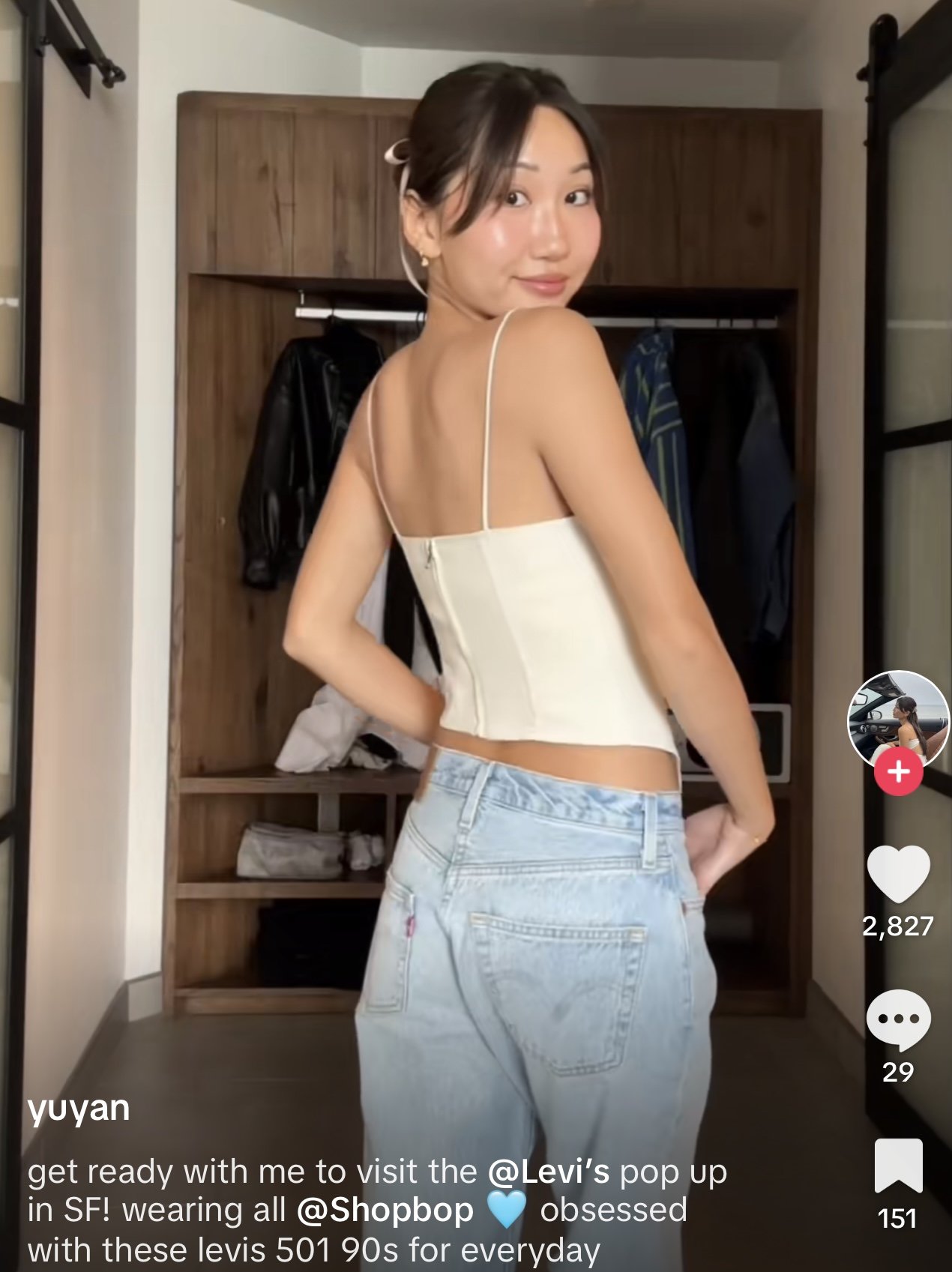 A young woman wearing a beige top and light blue Levi's jeans with a high waist standing in a room with a wooden closet in the background. She is smiling and looking over her shoulder at the camera.