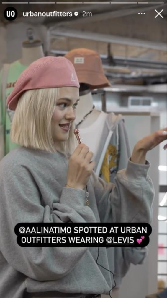 A woman with blonde hair wearing a pink beanie and gray sweatshirt, holding a lollipop, standing inside Urban Outfitters store with mannequins and clothing in the background.