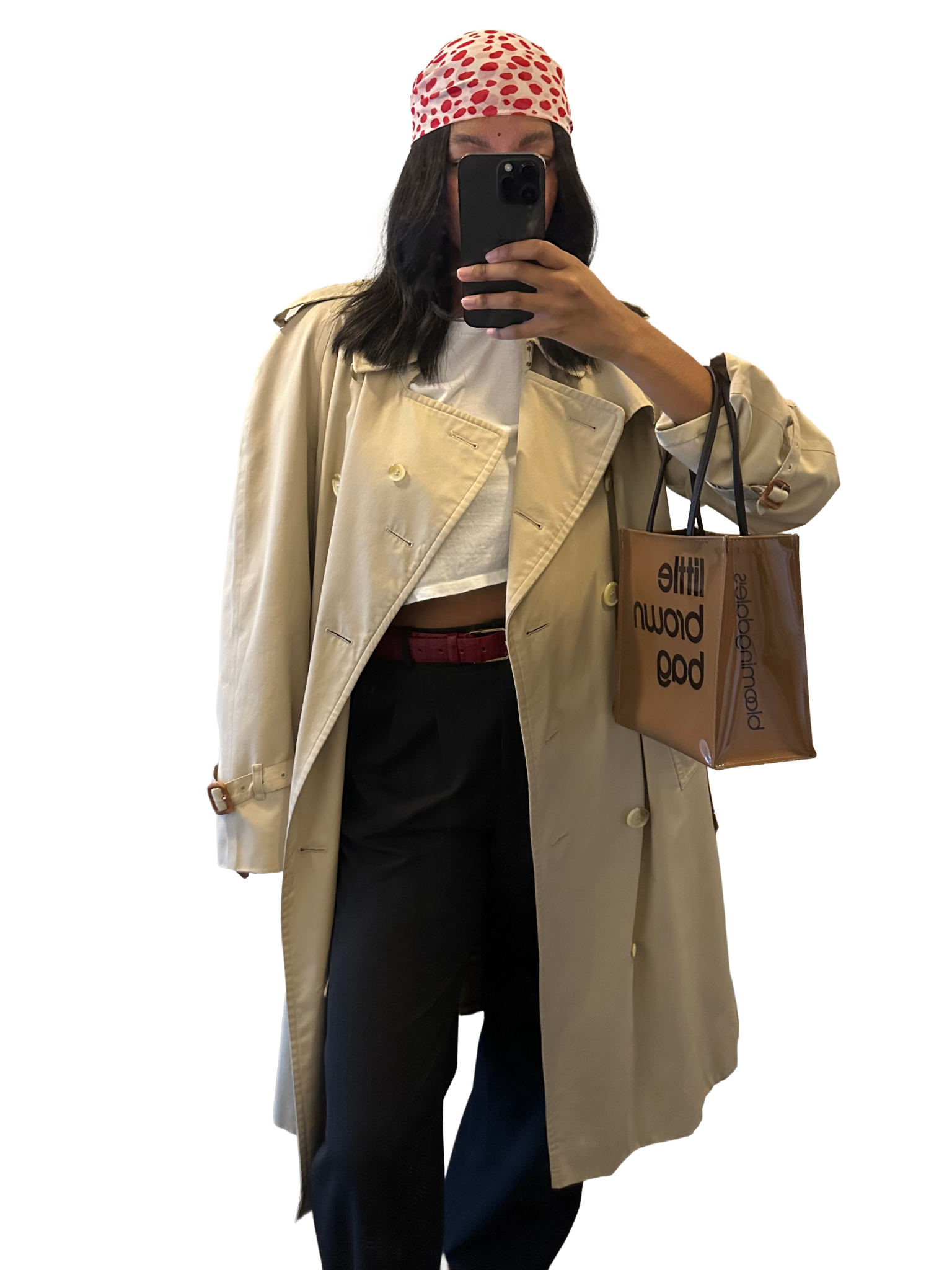 Woman taking a mirror selfie with a black phone, wearing a beige trench coat, white crop top, black pants, red belt, and a red polka dot headscarf, holding a brown shopping bag.