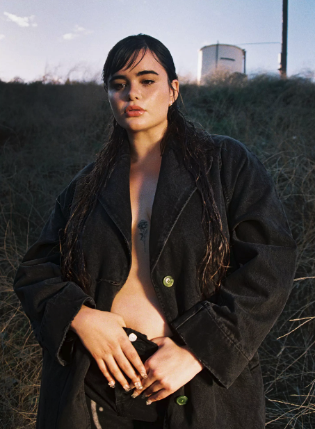 A woman with long dark hair and tattoos, wearing an oversized black denim jacket, standing outdoors with grass and a building in the background.