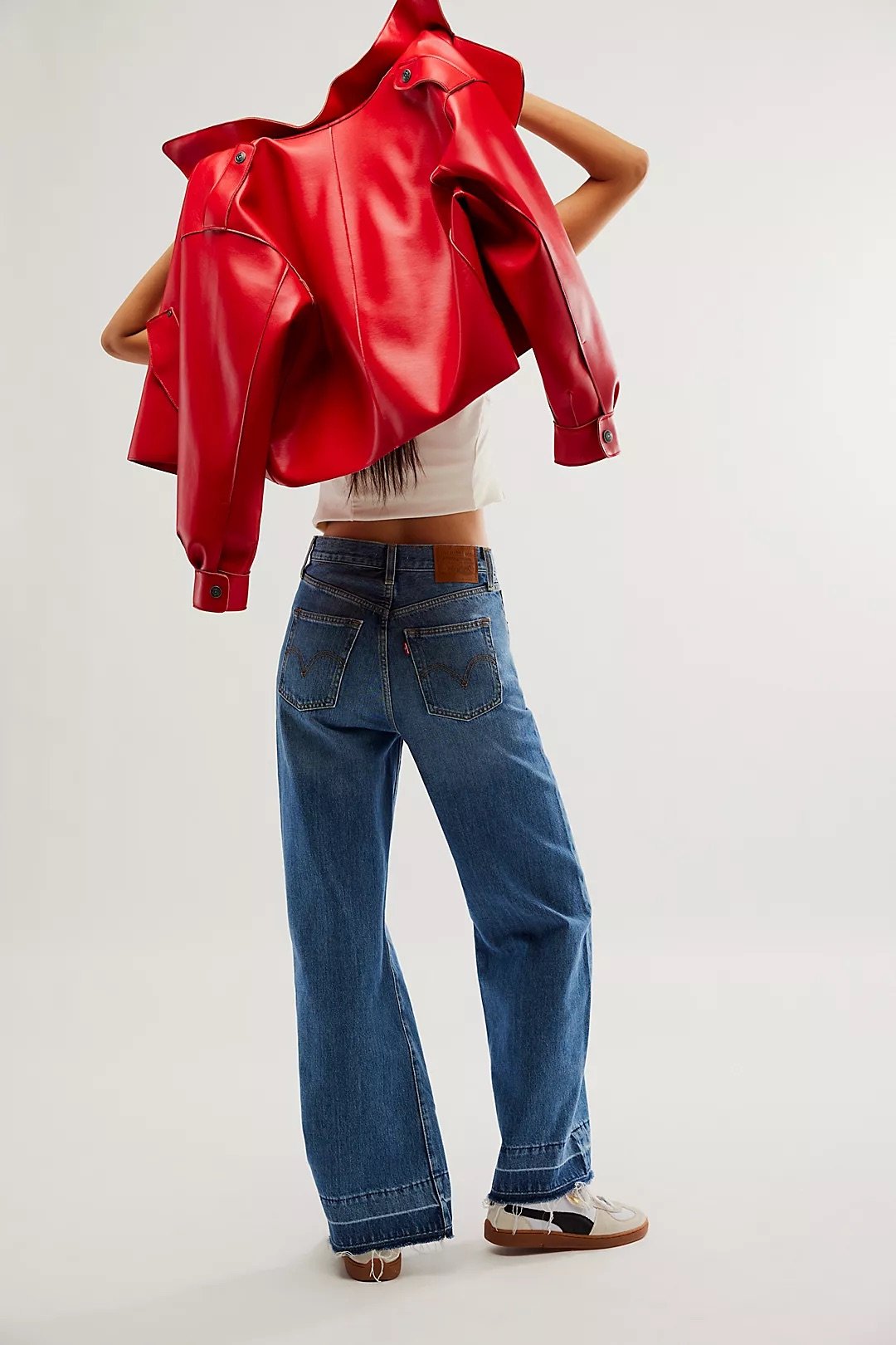 Person holding a red leather jacket over shoulders, wearing a white crop top, blue jeans, and white sneakers, standing against a plain neutral background.