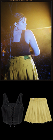 A woman with long hair wearing a black sleeveless top and yellow skirt, illuminated by stage lights.