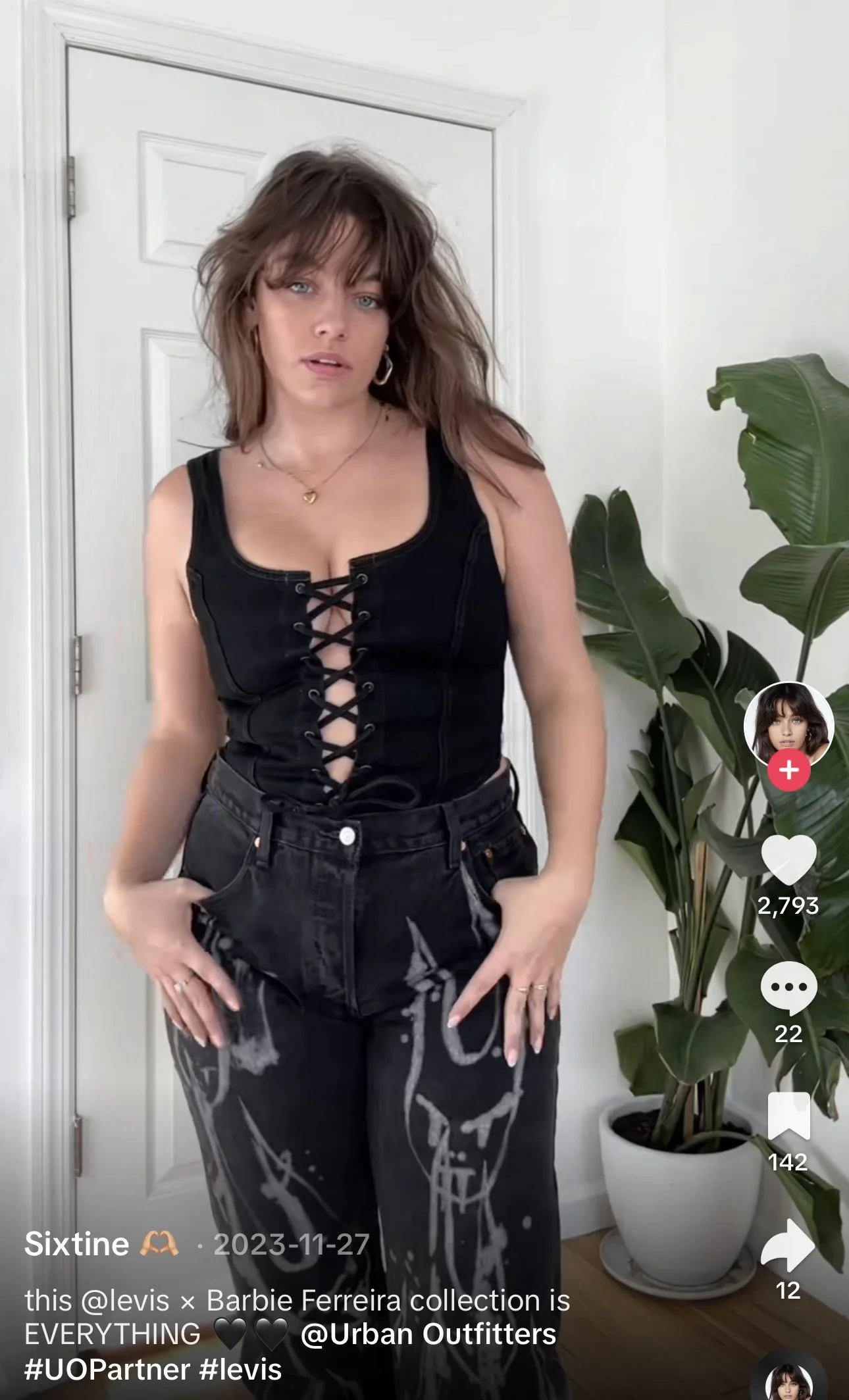 A woman with shoulder-length brown hair, wearing a black sleeveless top with a lace-up front and black pants with white graffiti-style writing, standing in front of a white door and a potted plant.