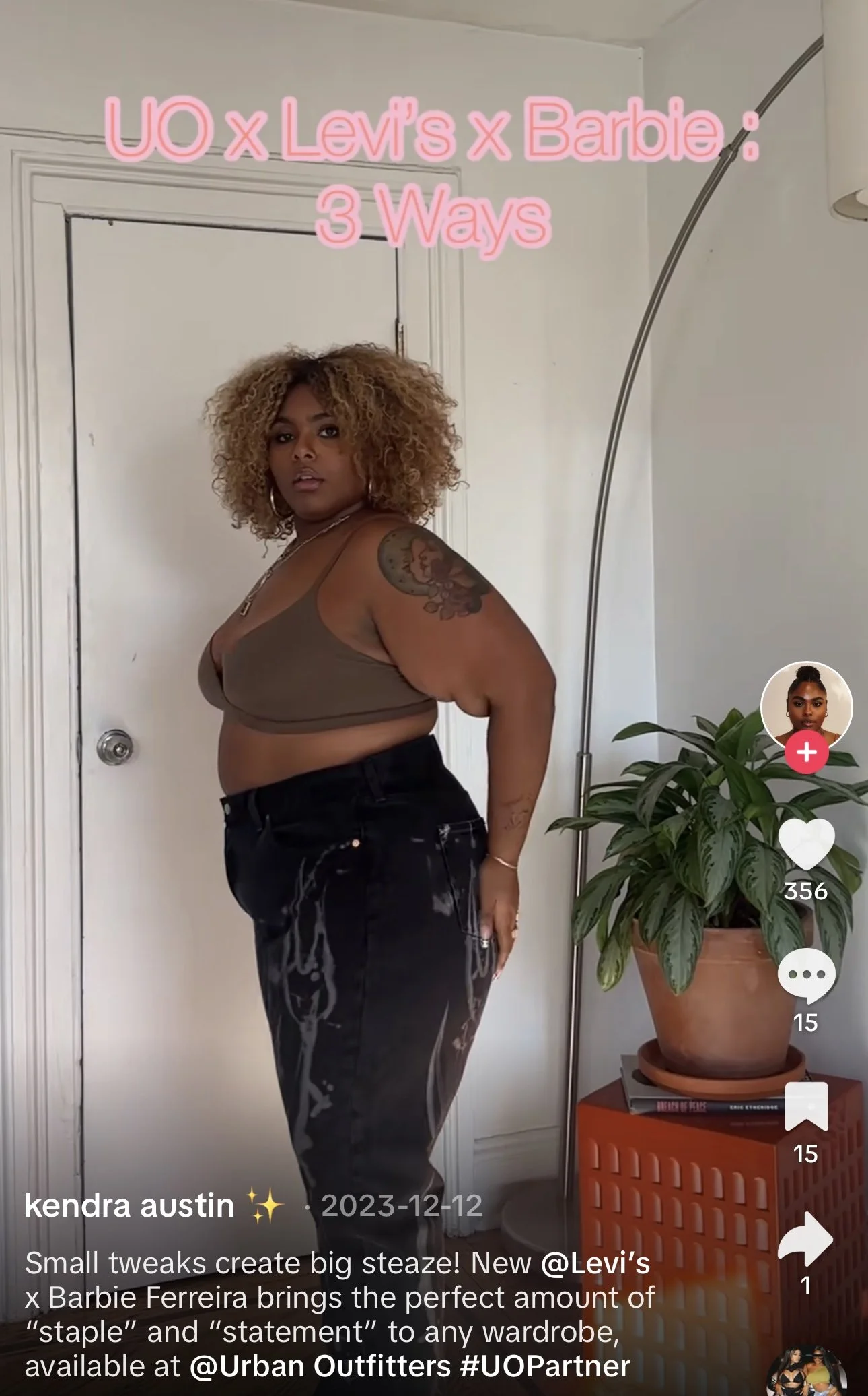 A woman with curly blonde hair and a tattoo on her right arm stands in front of a white door with a doorknob. She is wearing a brown tank top and black pants with a satin finish. To her right, there is a tall, curved silver floor lamp and a potted plant on an orange side table with books underneath. Pink text at the top of the image reads, 'UO x Levi's x Barbie: 3 Ways'.