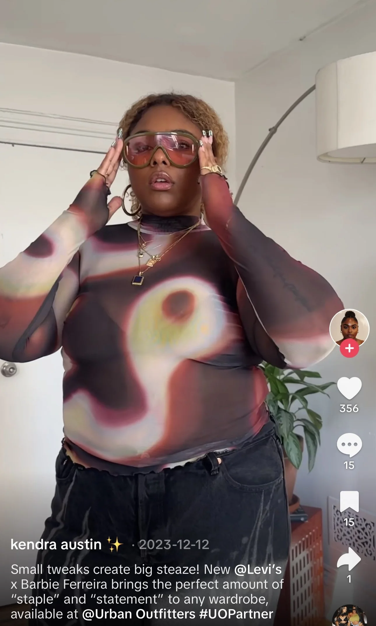 Woman with curly hair wearing pink-tinted sunglasses, black long-sleeve top with a colorful abstract pattern, and black pants, adjusting her glasses in a room with a lamp and a plant.