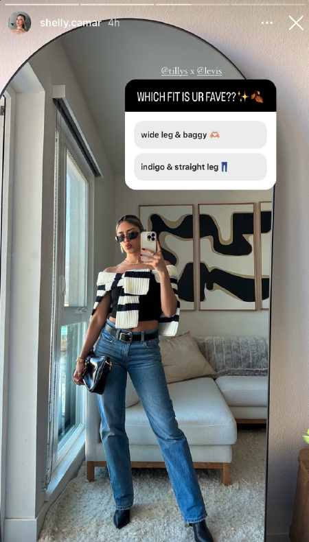 Woman taking a mirror selfie in a living room, wearing sunglasses, a black and white striped off-shoulder top, high-waisted jeans, and black boots, with a handbag on her wrist, and abstract artwork on the wall behind her.