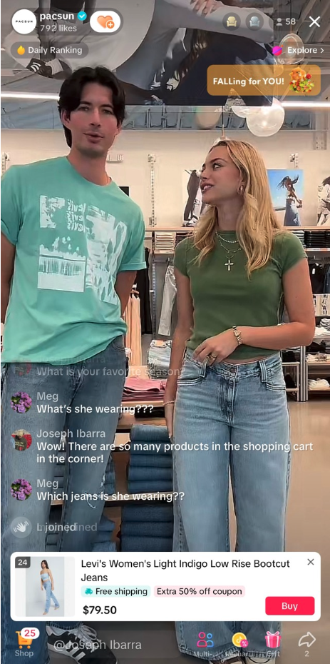 A man and a woman standing inside a clothing store. The man is wearing a light teal graphic t-shirt and jeans, while the woman is wearing a green short-sleeve top, jeans, and jewelry. They are engaged in a conversation, with clothing racks and shelves of folded clothes in the background.