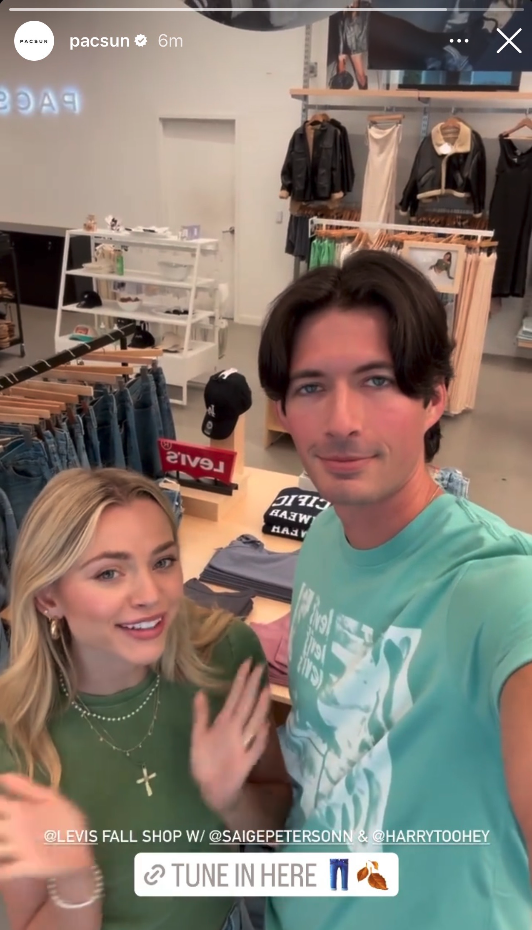 Two people taking a selfie inside a clothing store. The woman on the left has long blonde hair and is wearing a green T-shirt with jewelry. The man on the right has dark hair and is wearing a light turquoise T-shirt. Behind them are racks of jeans and various clothing items.