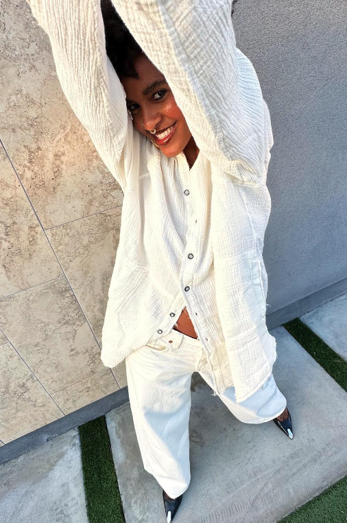 Woman smiling, wearing a white outfit, standing outdoors near a stone wall and gray concrete ground with small patches of artificial grass.