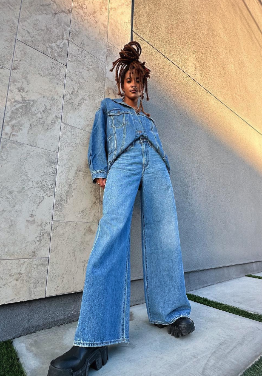 Young woman with dreadlocks wearing a denim jacket and wide-leg jeans, standing against a modern stone wall, looking at the camera.