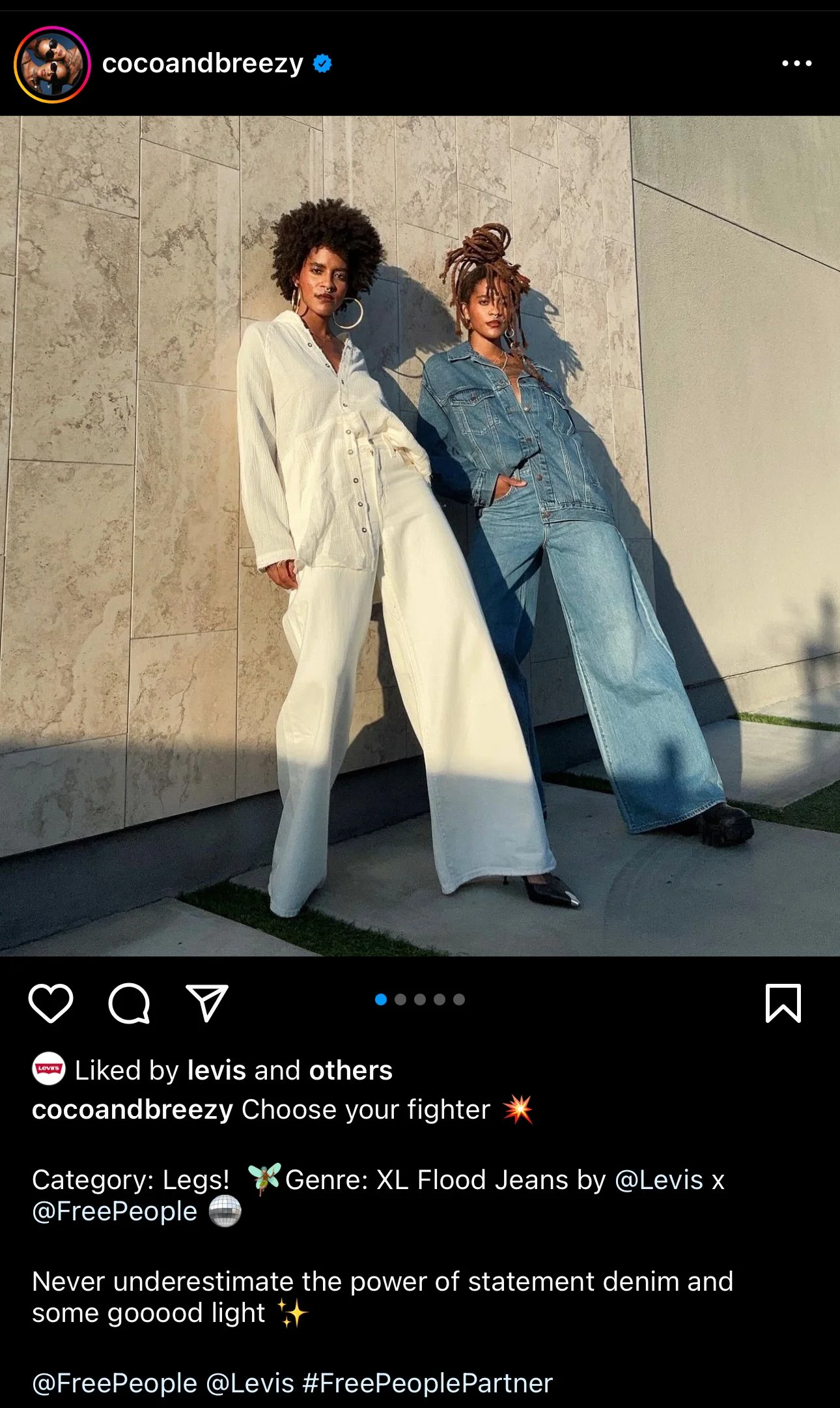 Two women standing against a stone wall, one wearing a white shirt and wide-legged jeans, the other wearing a denim jacket and wide-legged jeans, both with stylish hair and accessories, posing confidently.