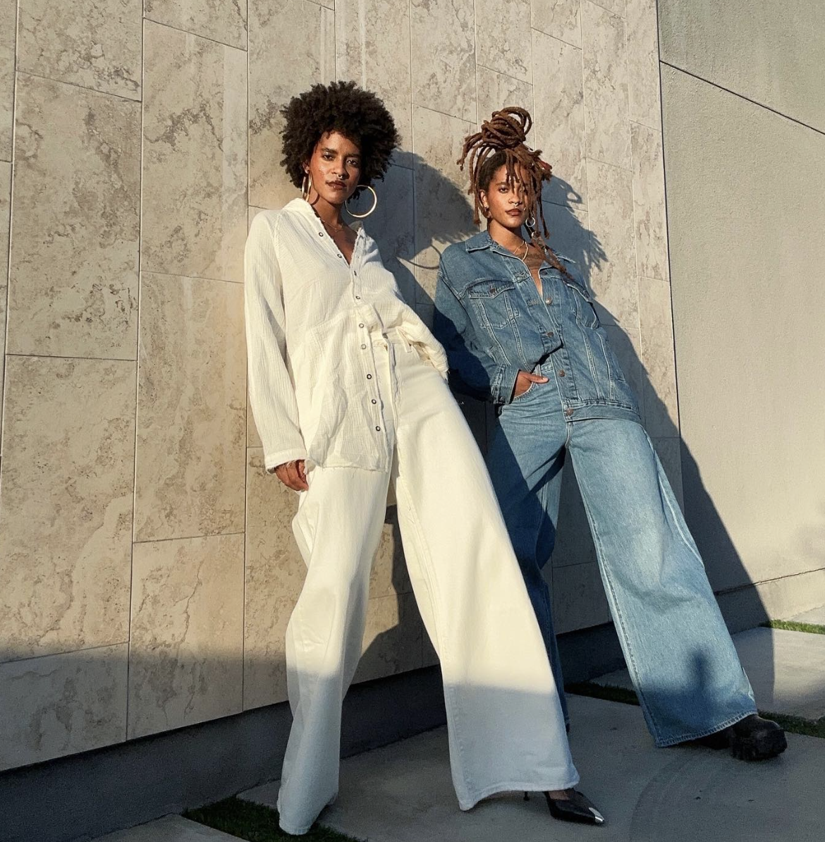 Two women with dreadlocks and hoop earrings leaning against a tiled wall, wearing stylish denim and white outfits, with sunlight casting shadows.