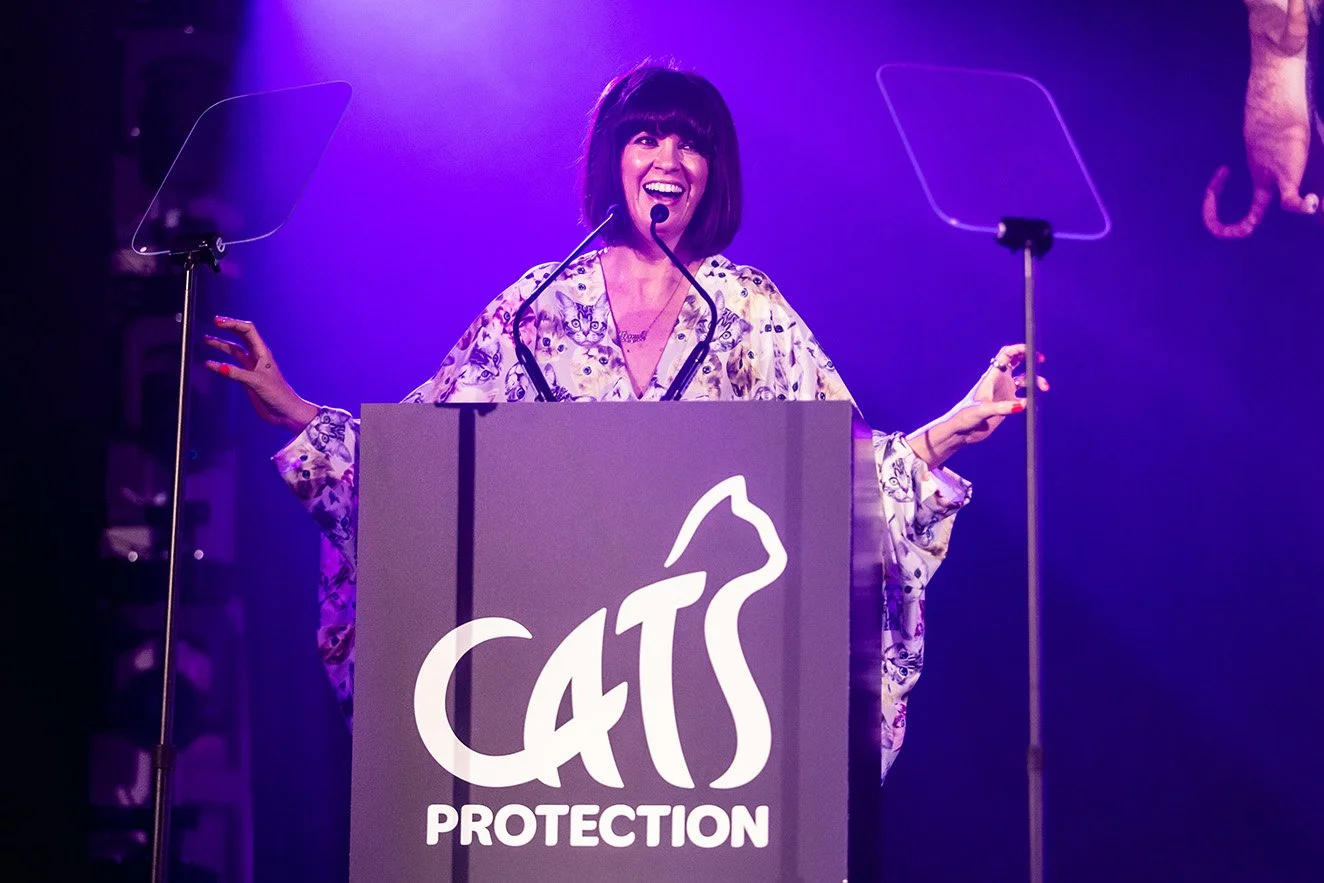A woman with dark hair and a short bob cut speaking at a podium with a sign that reads "Cats Protection." She is smiling and wearing a patterned blouse, with purple lighting in the background.